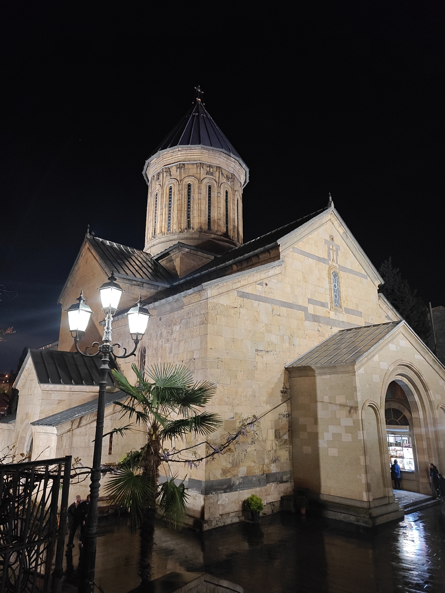 This nocturnal image showcases a prominent Georgian Orthodox church in Tbilisi, Georgia, bathed in warm light against a dark sky. The church features a striking central dome topped with a cross, constructed from light-colored stone blocks with decorative darker banding. The dome itself has numerous tall, narrow arched windows. The main body of the church also exhibits stone masonry, gabled roofs, and an ornate, tall, narrow window with a cross design on the upper wall of the right facade.
In the foreground, an elaborate black street lamp with three bright lanterns illuminates the scene, casting reflections on the wet ground, which suggests recent rain. A medium-sized palm tree stands between the lamp and the church, its fronds catching the light. On the right side of the church, an arched entrance leads into a well-lit interior, where a person in a blue jacket is visible, seemingly looking at something or entering. Another individual is partially visible near this entrance. Farther to the left, behind a dark metal fence or railing, two more people are faintly discernible. Potted plants are placed near the church walls.
The wet ground beautifully reflects the artificial lights, creating a shimmering effect that enhances the atmosphere of the night scene. No discernible text is visible.