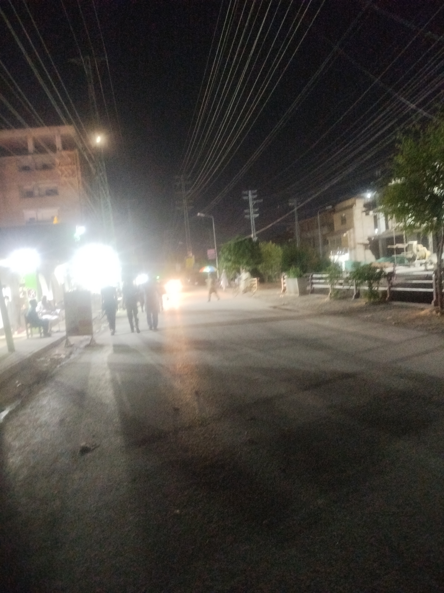 A nighttime, low-angle, slightly blurry, full shot of a street in Peshawar City Tehsil, Pakistan. A building with lights is visible on the left. A street with power lines stretching overhead is the main focus. Three figures, likely male, are walking on the right side of the street. Two more figures, possibly female, are on the far right, one holding an umbrella. Other individuals are seated on the left, near the building. Several parked vehicles, including a motorcycle and what appears to be a construction vehicle, are visible. The scene suggests a populated area with commercial activity.