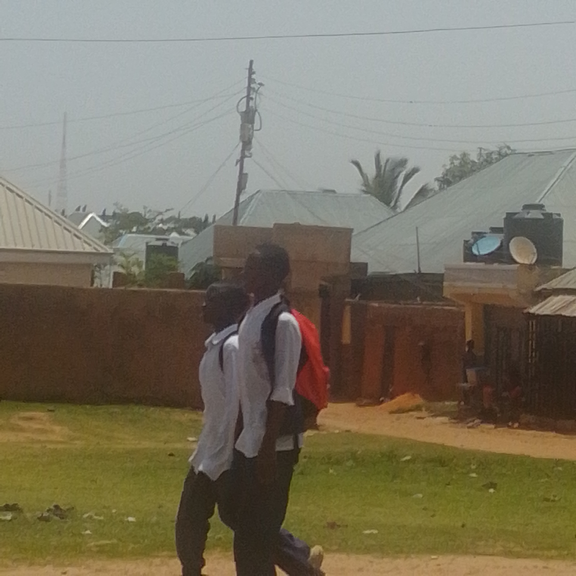 In Gombe, Nigeria, on a hazy day, two individuals wearing white shirts and dark pants walk side-by-side on a dirt path next to a grassy area. Both individuals have dark skin and black hair. The person on the left is slightly behind the person on the right. The person on the right has a red backpack slung over their left shoulder. In the background, there are several structures with light-colored, sloped roofs, and a brown wall. A power pole with wires is visible on the left, and a water tank and satellite dish are visible on a rooftop in the distance. The overall impression is of a residential or semi-urban environment.