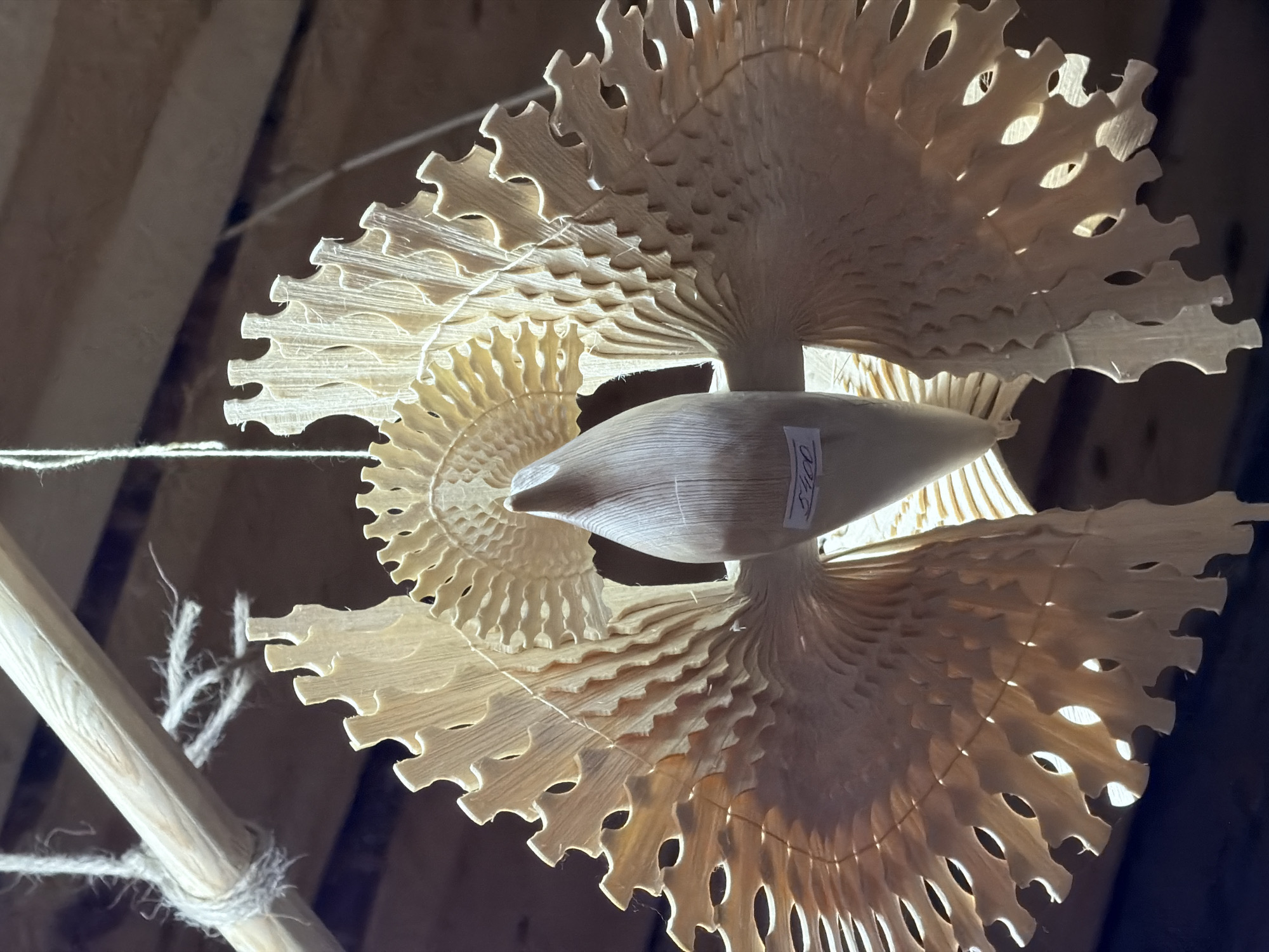 An ornate, handcrafted wooden bird sculpture hangs suspended from a wooden beam by twine. The bird has a smooth, conical body, and its wings are intricately carved from thin pieces of wood, creating a delicate, layered appearance with many small openings. The wings fan out in a semicircular shape, resembling feathers. A small white price tag, with the number "540" visible, is attached to the bird's body. The background consists of rough wooden ceiling planks. The sculpture appears to be in a workshop or craft store setting in the city of Imeni Sverdlova, Russia.