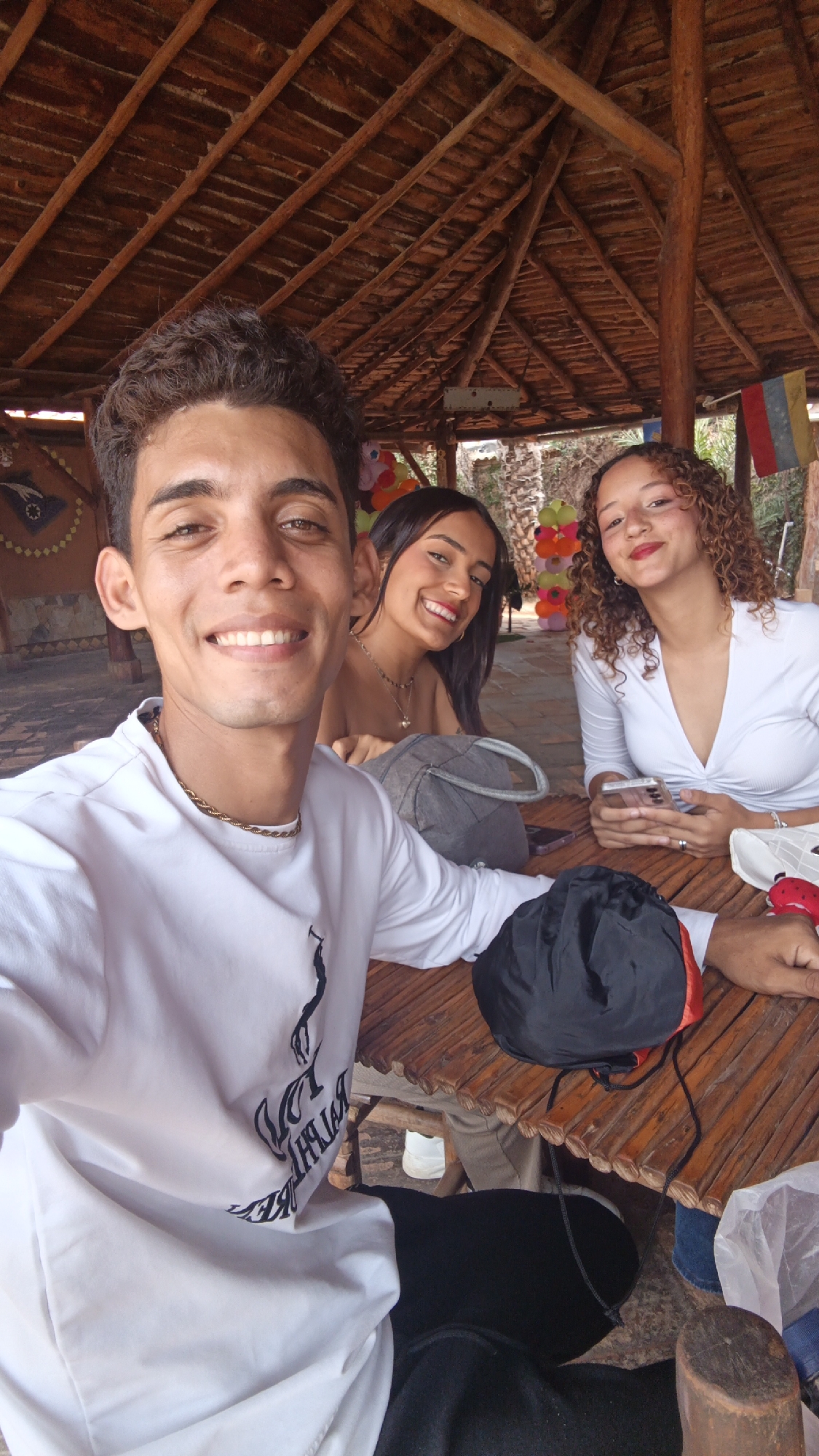Three young adults are seated at a rustic wooden table beneath a thatched-roof structure. A fair-skinned man in a white long-sleeved shirt with a dark graphic design is in the foreground, smiling at the camera, seemingly taking a selfie. He wears a gold chain. To his left is a fair-skinned young woman with long dark hair, also smiling. To his right is a fair-skinned young woman with curly reddish-brown hair, wearing a white top and holding a mobile phone. A black hat rests on the table, along with a grey tote bag. Behind them, decorations including balloons and a Venezuelan flag are visible. The structure appears to be open-air, and the location is likely a restaurant or gathering place in Juan Griego, Venezuela, evidenced by the flag and based on details in the background.