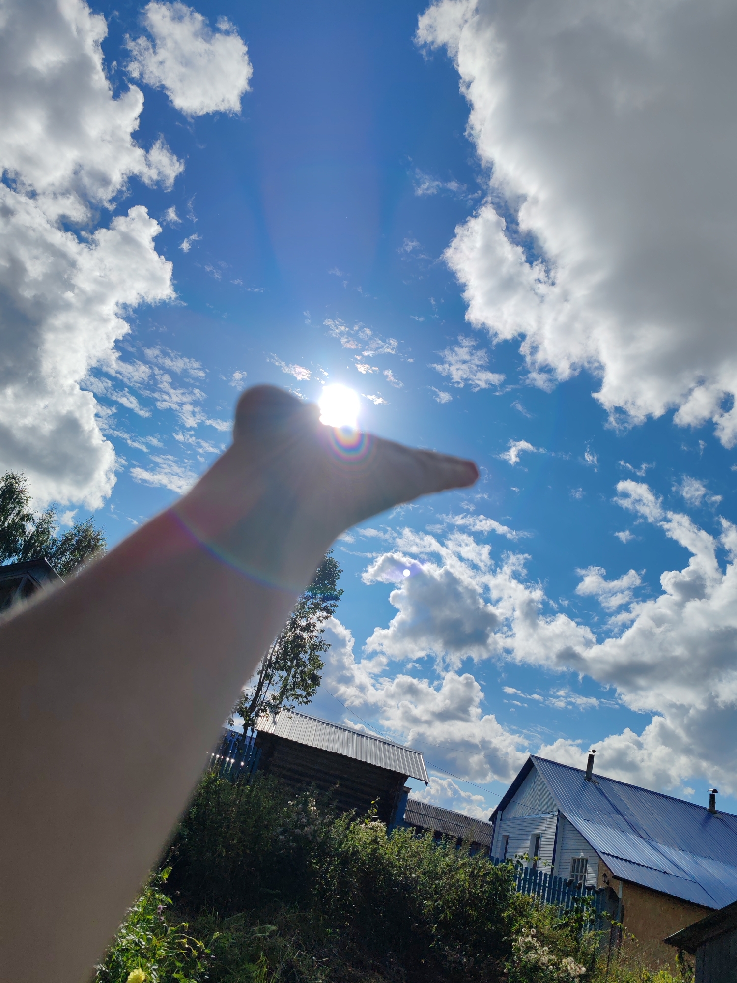 In Чухлэм, Russia, on a bright, sunny day, a hand is held up towards the sky, partially obscuring the sun. The sun, a brilliant white disc, creates a lens flare with rainbow hues. The sky is a vibrant blue, dotted with fluffy white clouds. Below the sky, a wooden building with a corrugated metal roof is visible, along with green trees and foliage. A blue wooden fence runs in front of another building with a blue metal roof and a chimney. The scene appears to be a rural or suburban setting.
