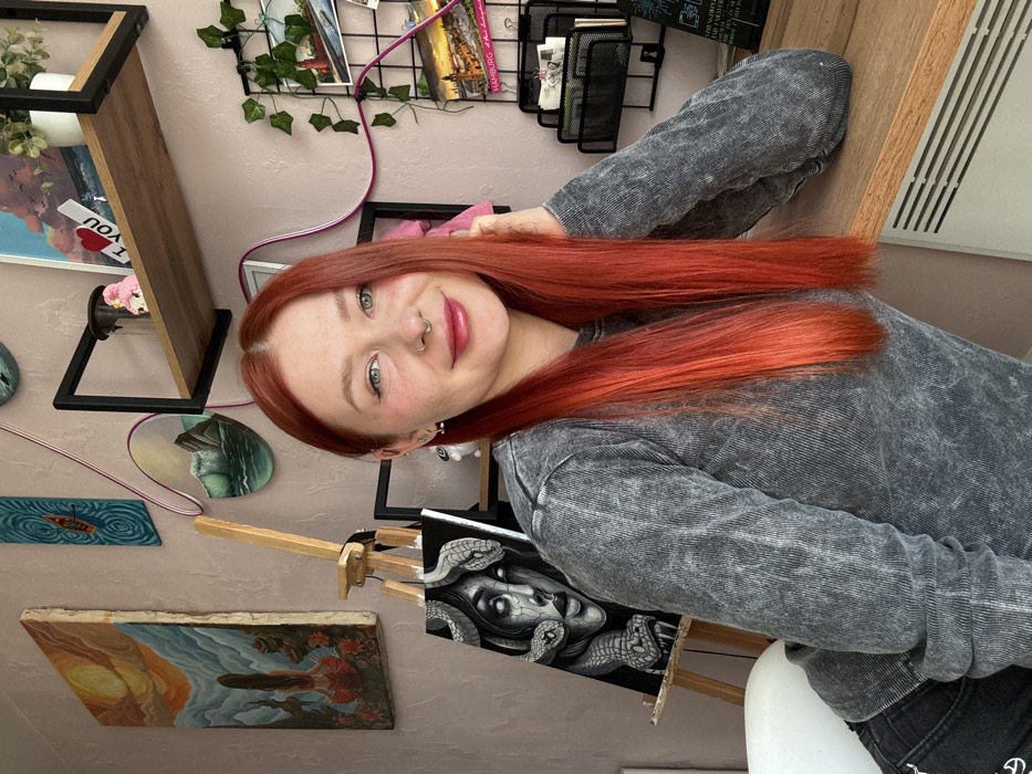The image displays an indoor scene in Rostov-on-Don, Russia, featuring a young adult female subject. The woman, positioned centrally, has long, straight, vibrant red hair and light-colored eyes. She exhibits a septum piercing in her nose and small stud earrings in both earlobes. Her attire includes a long-sleeved, grey, acid-washed ribbed top and dark pants with a white graphic on the left thigh, partially visible as "CHAMPION". She is leaning on a light-colored wooden desk or table with her right elbow, looking directly at the camera with a slight smile.
The background wall is a uniform light pinkish-beige. To the left, a wooden easel holds a black canvas displaying a greyscale painting of a woman with snakes for hair. Above and to the left of the easel, a large rectangular painting of a figure by a sunset and a circular painting of a turbulent sea are mounted. A black metal and wooden shelf above the subject's right shoulder holds a small potted plant, a pink figurine, and a framed artwork with an "I LOVE YOU" sign. A black metal grid board to the right features artificial green vines, several postcards (one identifiable as "HAMBURG"), and a black mesh organizer containing papers. Pink electrical cords or decorative lines are visibly traversing the wall. A white slatted radiator is located beneath the desk. A white chair is partially visible on the far left.