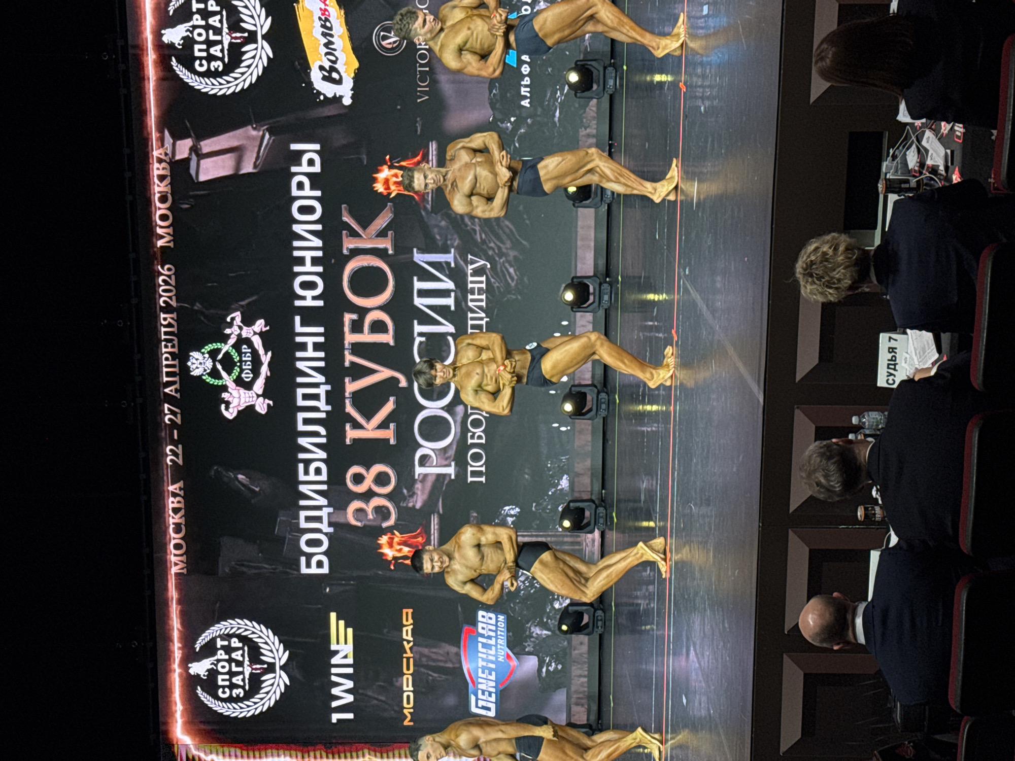 This image captures an indoor bodybuilding competition, likely taking place on a stage with a dark background. Four male bodybuilders are visible on the stage, all in posing trunks, displaying well-defined musculature and a deep tan. They are performing various poses, facing an unseen audience or judges. Bright stage lights illuminate them from the floor.
Behind the bodybuilders, a large banner serves as a backdrop, featuring event information and sponsor logos. The banner clearly indicates the event as "БОДИБИЛДИНГ ЮНИОРЫ" (Bodybuilding Juniors) and "38 КУБОК РОССИИ" (38th Russian Cup). It also specifies the location and dates: "МОСКВА 22 - 27 АПРЕЛЯ 2026" (MOSCOW 22 - 27 APRIL 2026). Prominent logos on the banner include "СПОРТ ЗАГАР" (Sport Tan), "ФББР" (FBBR - Federation of Bodybuilding and Fitness Russia), "1 WINE", "МОРСКАЯ", "GENETICLAB NUTRITION", "BOMBBA", "VICTORC", and "АЛЬФА".
In the foreground and to the right of the stage, several individuals are seated at tables, presumably judges or officials. Their backs are mostly visible, and one person wears a badge or tag that reads "СУДЬЯ 7" (Judge 7). They appear to be observing the competition intently, with papers and water bottles on their tables. The overall atmosphere suggests a focused and professional sporting event.