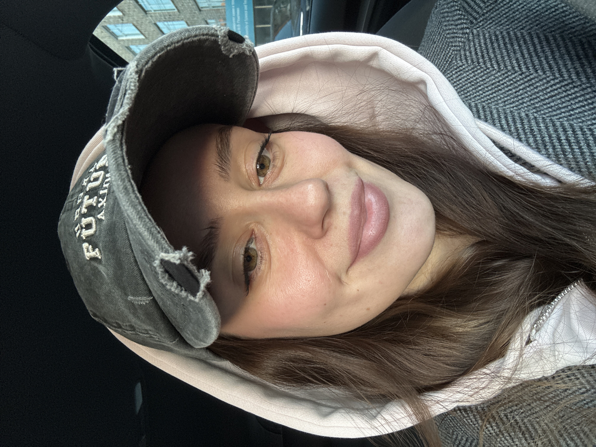 A young woman wearing a worn, dark grey baseball cap with a light pink hoodie underneath a grey herringbone jacket is taking a selfie. Part of a building can be seen through the vehicle window in the upper left corner. It is not possible to determine the exact location in Krasnoyarsk, Russia from the image.