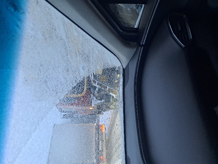 The image depicts a vehicle's interior, likely the driver's side. A frosty windshield obstructs much of the outside view, showing glimpses of other vehicles, including a truck, and what appears to be a snowy or icy landscape. Interior details include the dashboard, a window, and an air vent. The view is partially obscured by the frosty conditions on the windshield.