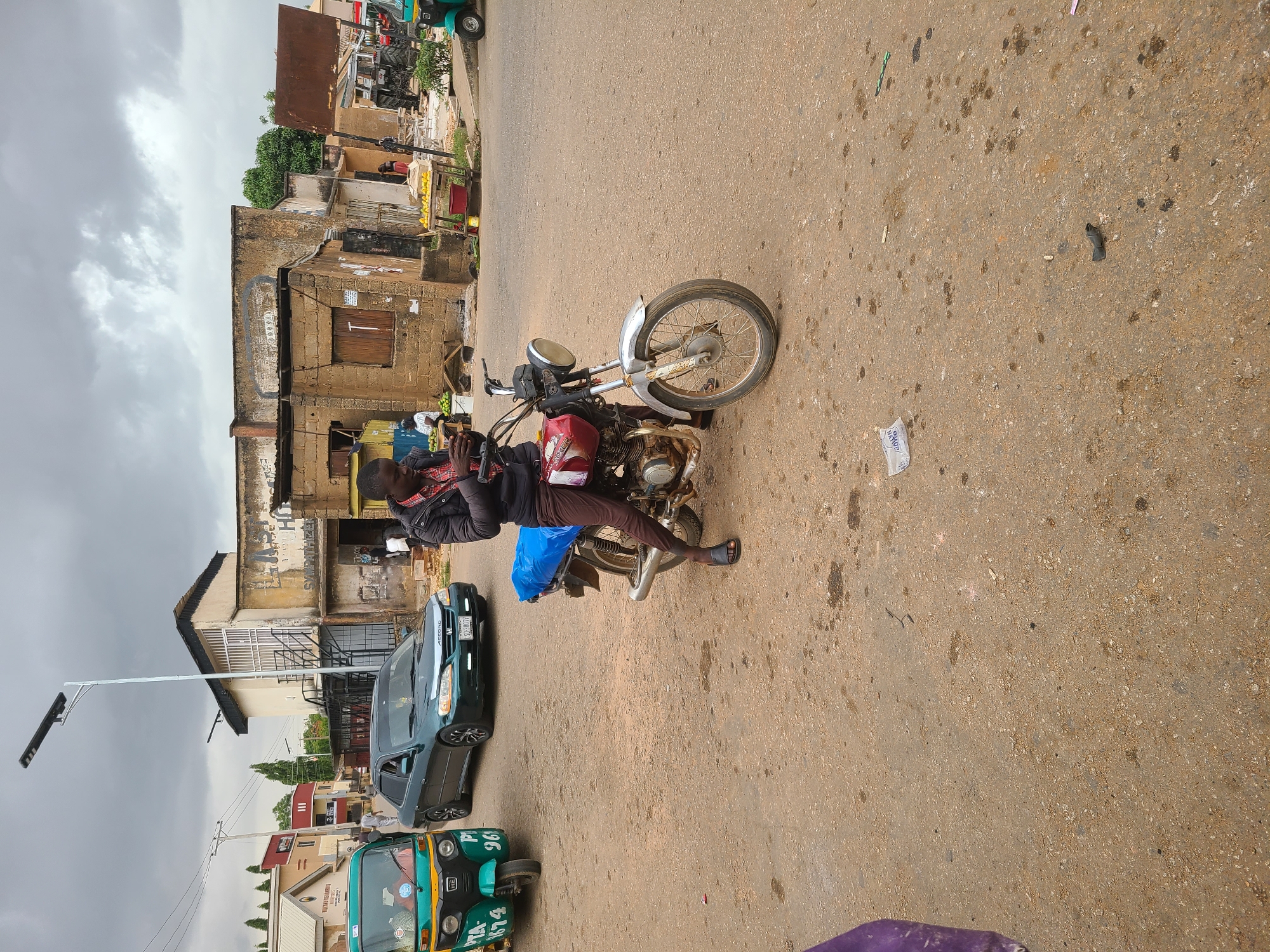 A man is sitting on a motorcycle on a paved street in Gura Top, Nigeria. He is wearing a dark jacket and brown pants. A blue bag rests on the motorcycle behind him. The street is lined with various vehicles, including a green tricycle, and a dark green car. Buildings in varying states of repair stand on the side of the street. A street lamp is visible in the foreground. The sky is overcast.