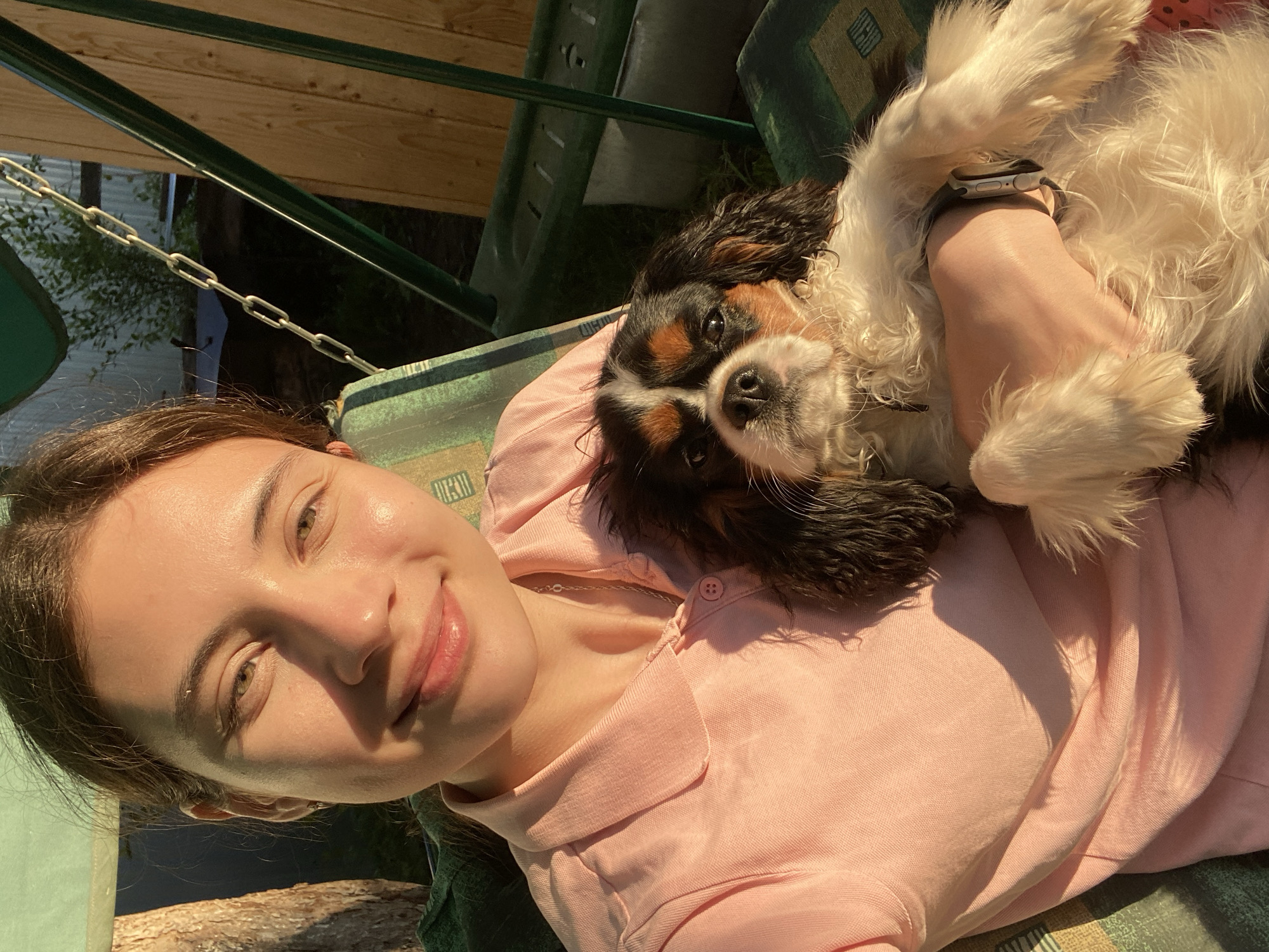 In an outdoor setting, possibly a backyard in Votkinsk, Russia, a young woman with brown hair pulled back is holding a Cavalier King Charles Spaniel dog. She is wearing a light pink collared shirt and a silver necklace. The dog has a tricolor coat and is looking directly at the camera. They are both seated on a green swing, which has patterned cushions. The late afternoon sun casts warm light and shadows on the subjects and the surrounding environment, which includes wooden fencing and vegetation.