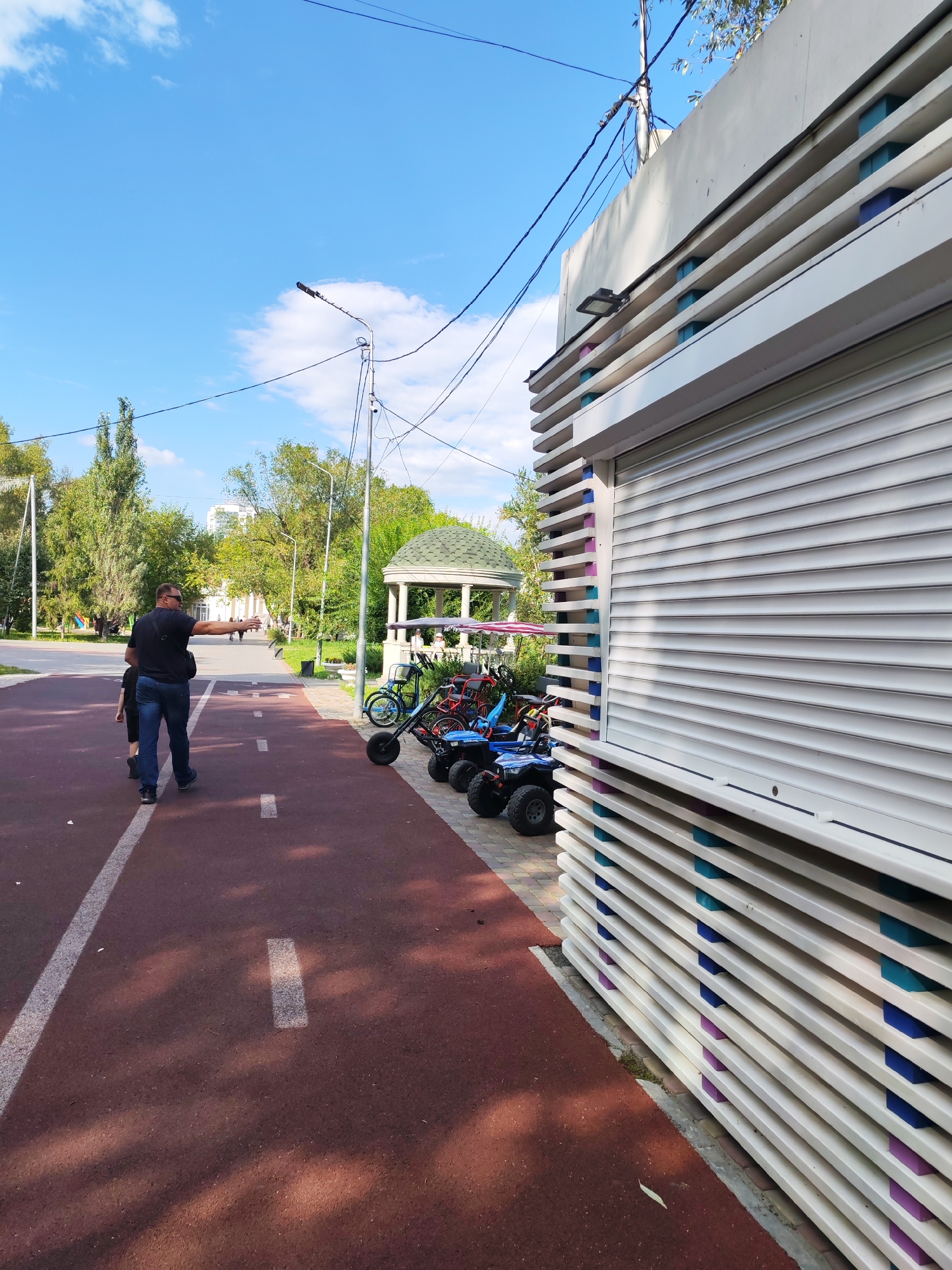 In an outdoor park setting, a man in a dark t-shirt and jeans, wearing sunglasses, is walking away from the camera on a red paved path marked with white lines. He is gesturing with his right hand. A child, also walking away, is ahead of him. To the right of the path are several small vehicles resembling go-karts or quad bikes, some blue and some red. A white building with horizontal louvers and colorful blocks (blue and purple) behind them is situated next to the vehicles. In the background, there are trees, a gazebo with a green roof, and a clear blue sky with scattered clouds. The overall scene depicts a recreational area on a sunny day.