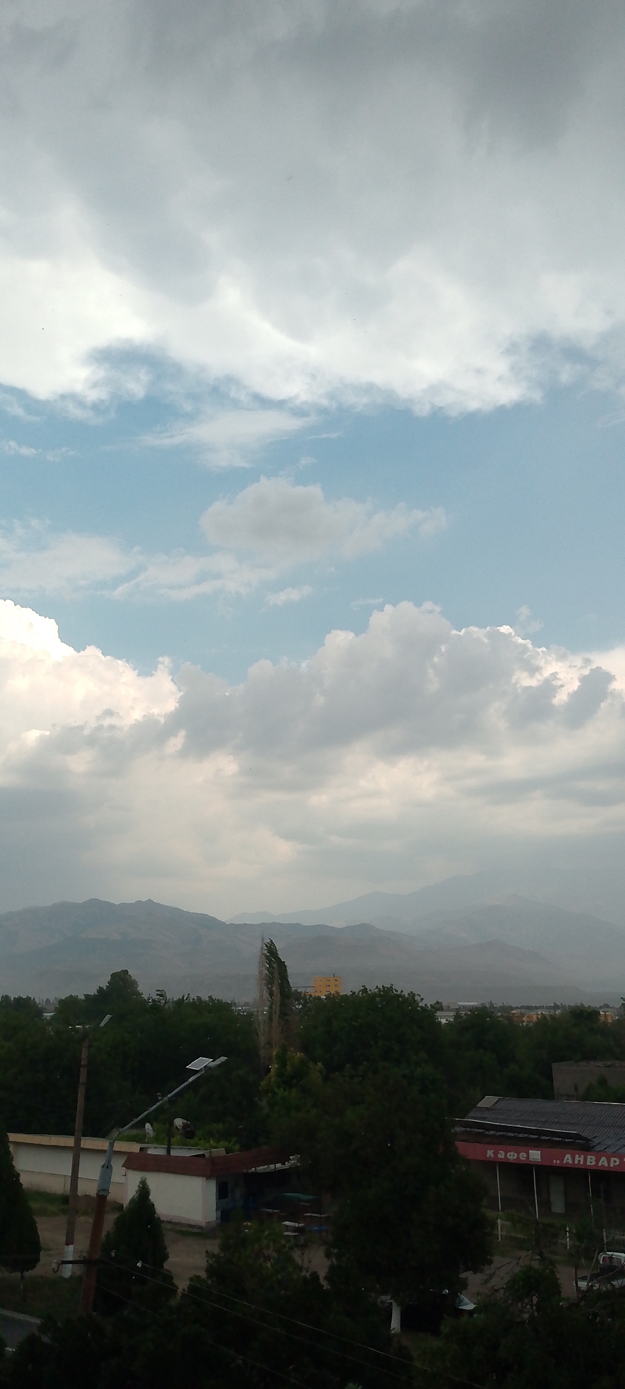 The image depicts a landscape view in the city of Angren, Uzbekistan, under a partially cloudy sky. In the foreground, dense green foliage of trees dominates the lower half of the frame. Several small buildings are visible amidst the trees, including a structure with a red awning and Cyrillic lettering that appears to read "КАФЕ, АНВАР". A street lamp with solar panels is also present. In the mid-ground, a tall, thin, monument-like structure is positioned against a backdrop of trees and a distant yellow building. The background features a range of hazy mountains. The overall scene suggests a semi-urban or suburban environment with natural elements.