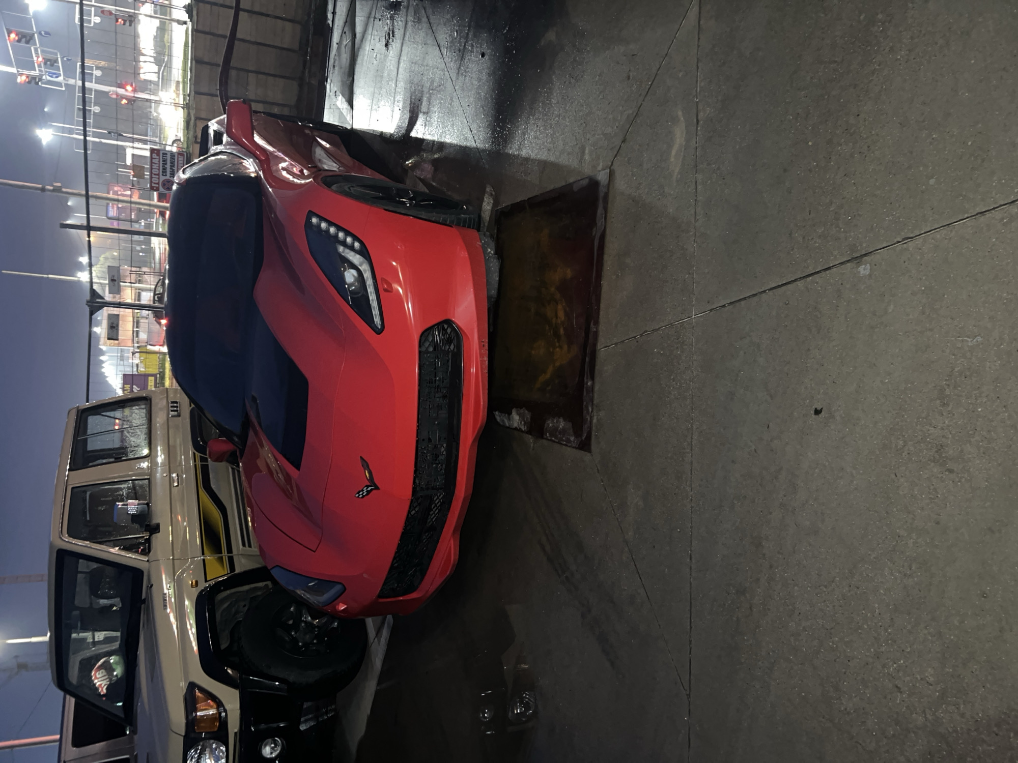 The image shows a red sports car parked next to a beige SUV-style vehicle on a paved surface. The car is positioned over a rectangular, metal grate or drain. Traffic lights and streetlights are visible in the background, suggesting an urban setting. Based on the presence of a sign in Bulgarian, the location is Plovdiv, Bulgaria. The sky is dark, indicating it is likely nighttime.
