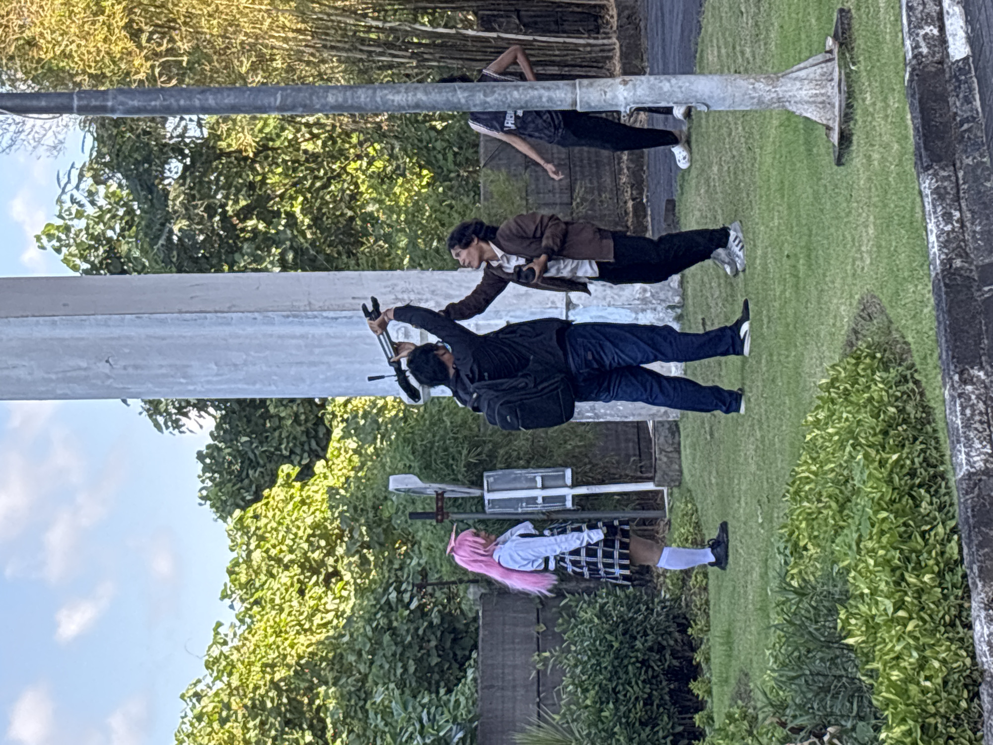In Kuta, Indonesia, four individuals are visible on a lawn in front of lush green foliage and bamboo. Two men in the foreground are actively engaged in manipulating a tripod, suggesting a filming or photography session. One man, wearing a dark jacket and jeans, holds the top of the tripod, while the other, in a brown jacket, holds a camera. To the left, a person with bright pink hair, dressed in a white shirt and a plaid skirt, stands looking away from the camera. Further to the right, another individual is partially visible, wearing a dark tank top and black pants. The scene is illuminated by natural daylight, with a clear sky overhead. A tall, cylindrical white structure stands prominently in the background, likely a pole or column.