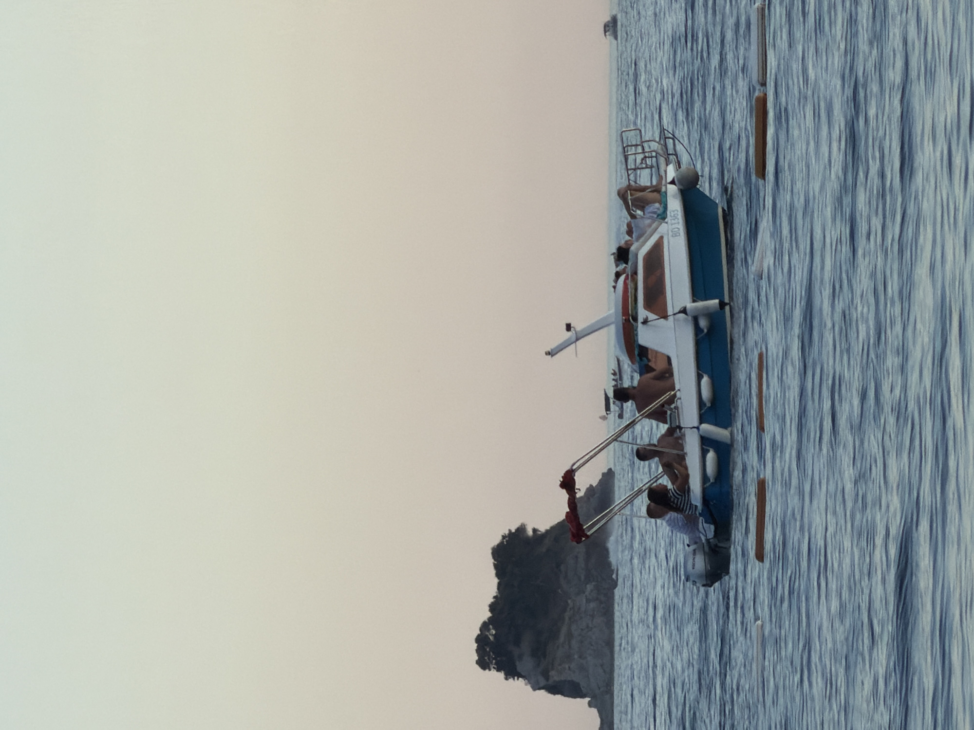 A small motorboat carrying at least five people is cruising on the calm, blue sea. The boat has a white and blue hull, a red canopy, and a Honda outboard motor. It is heading towards the right of the frame. Several people are seated or lounging on the boat. In the background, a rocky island covered in dark green foliage is visible to the left. The sky is a pale, hazy gradient from light blue at the top to a peachy-pink near the horizon. A few buoys are floating in the water. In the far background, another small boat is visible on the horizon. The overall scene suggests a leisurely day at sea near Budva, Montenegro.
