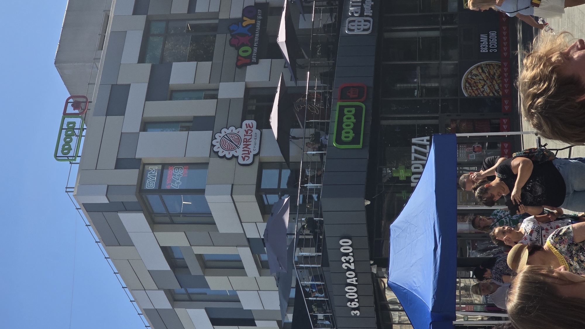 The image captures a commercial building on a sunny day in the city of Zhytomyr, Ukraine. The building features modern architecture with a facade of alternating grey and white rectangular panels. Prominently displayed on the building are various signs for businesses: "FORA" (a grocery store chain) and "GYM" are visible on upper levels, along with a sign for "SUNRISE" sushi bar and "YOYO" children's entertainment center. Below, a "FORA" sign is also visible, as is a sign for "PIZZA". The ground level shows a street scene with a blue canopy tent set up, under which several people are gathered. Some individuals are looking at their phones, while others are engaged in conversation. The overall impression is of a bustling commercial area.