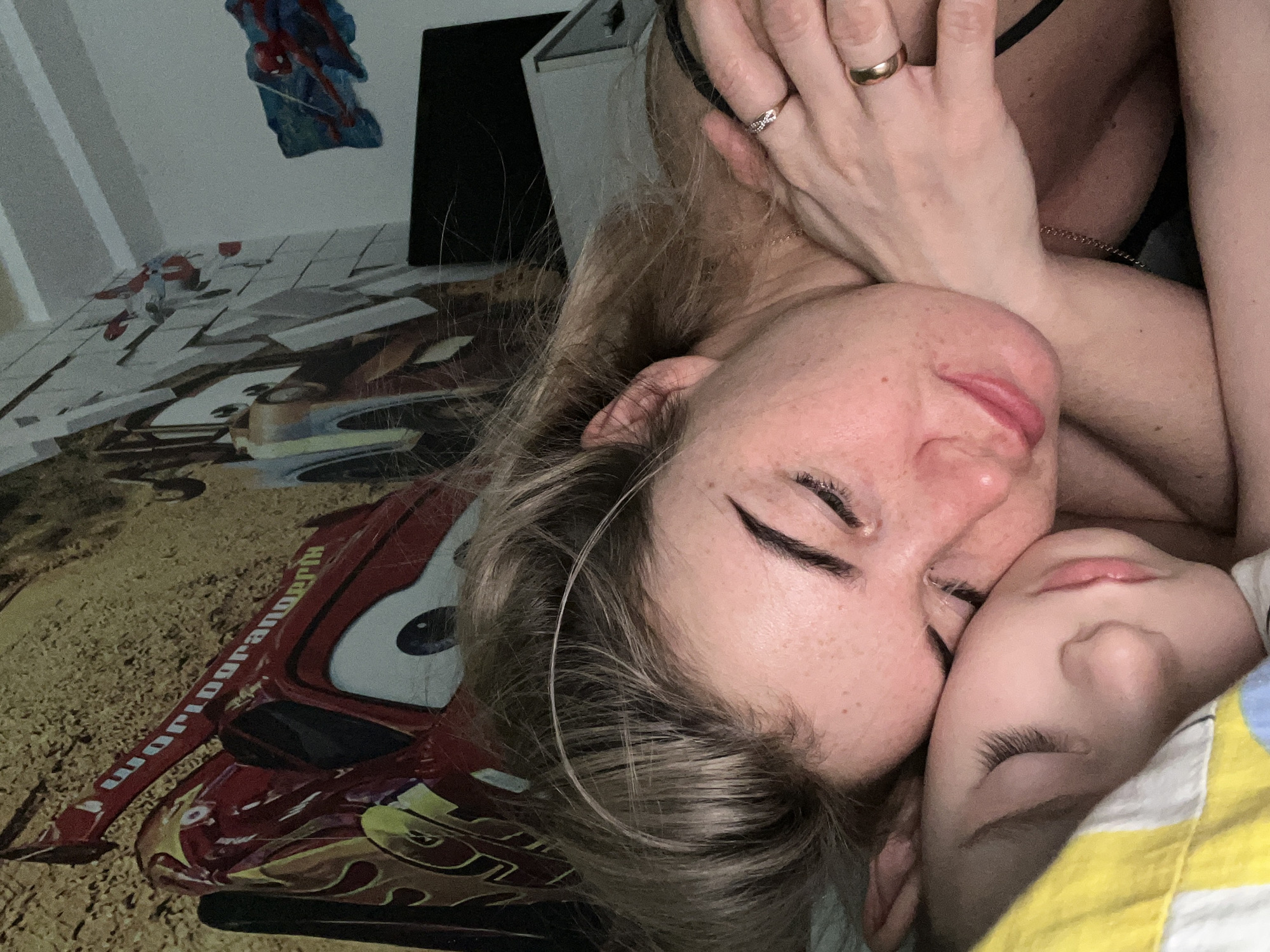 A close-up, high-angle portrait captures an adult woman and a child resting together, likely asleep, in an indoor setting in Chaikovsky, Russia. The image is dimly lit, suggesting evening, night, or early morning, and conveys a sense of peace and intimacy.
The adult, with light-colored hair and visible freckles on her face, lies on the left side of the frame, her eyes closed. Her left hand, adorned with a diamond ring on her ring finger and two gold bands, rests near her head. Cuddled closely beside her on the right is a young child, also with closed eyes, sleeping soundly and partly covered by a bright yellow and blue striped blanket.
Below the adult, a red bedspread or blanket features a large graphic of Lightning McQueen from Disney's Cars. Visible text on the side of the car includes a stylized or reversed phrase, possibly "wordgrandertk," consistent with character branding.
In the background, a light-colored wall with vertical stripes is visible, along with a blue and red Spiderman decal or poster. Some indistinct furniture or shelving can also be seen in the upper right background. The overall mood is one of quiet comfort and tender closeness between the two individuals.
