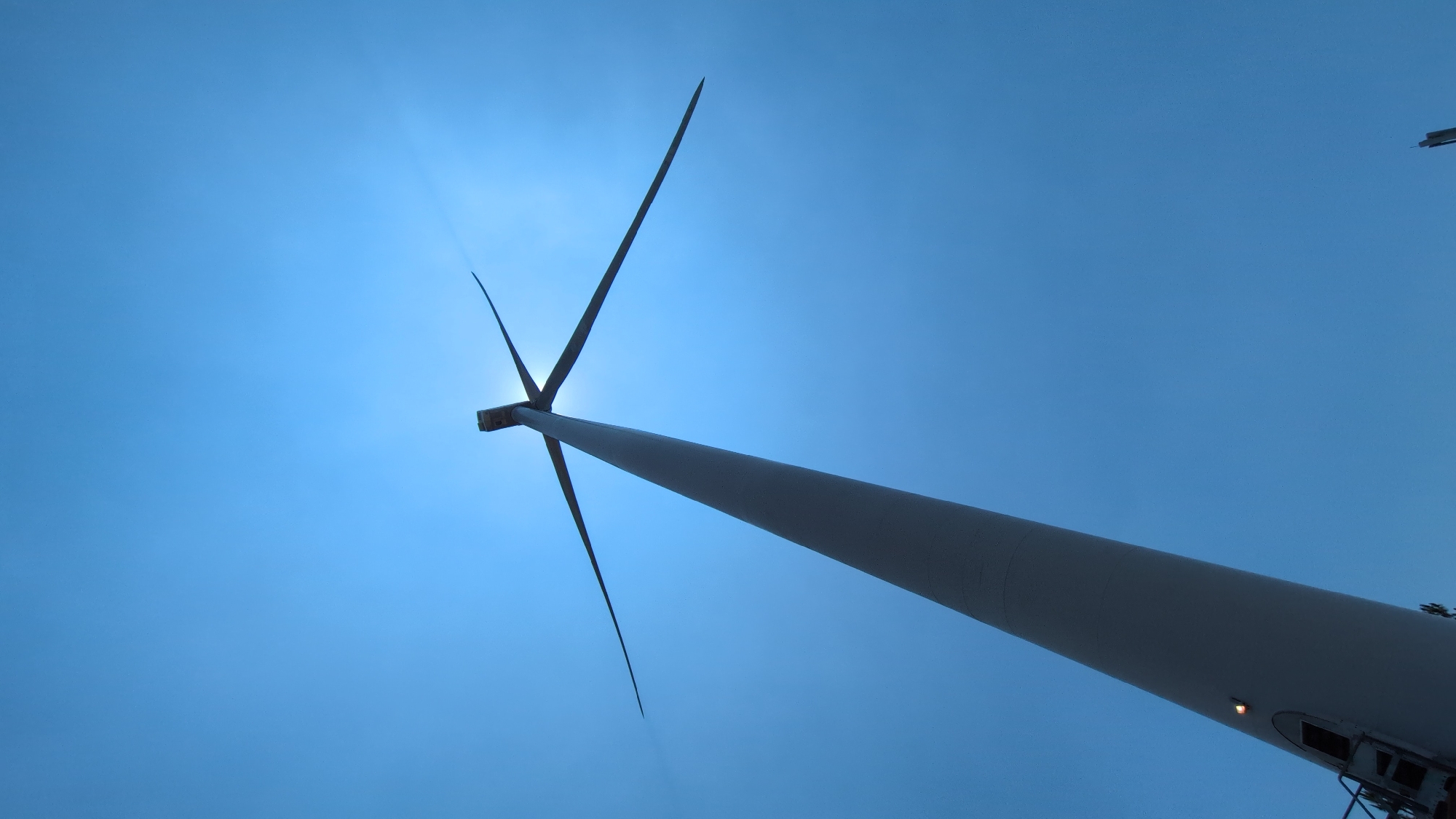 The image depicts a low-angle view of a wind turbine against a blue sky. The turbine's blades are silhouetted against the sun, which is directly behind the hub. The turbine's tower extends upward, angled towards the right side of the frame. The location is likely near Phát Chi, Vietnam, but that cannot be definitively determined from the image alone.