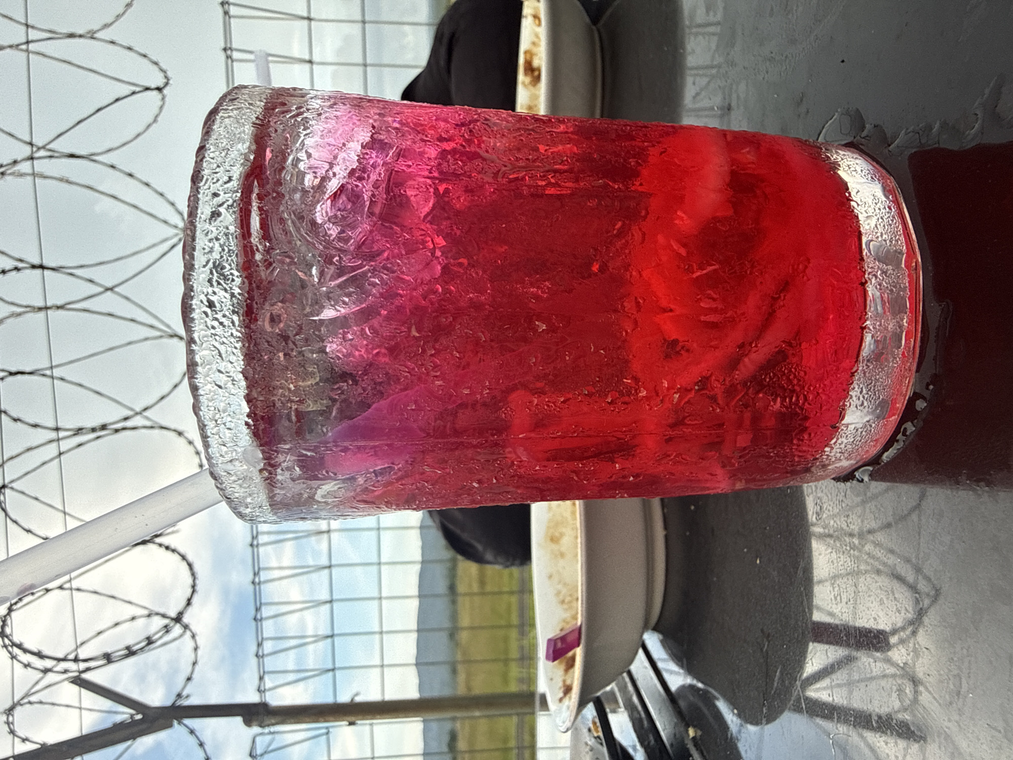 A glass of red, iced beverage sits on a dark, reflective surface. The glass is frosted with condensation. A white straw protrudes from the drink. In the background, a white bowl with a purple utensil is visible. Beyond that, a black object, possibly clothing, rests on a surface, and a wire fence or barrier with barbed wire runs across the top of the frame. The sky is cloudy, and a hilly landscape is visible in the distance. The scene appears to be outdoors, potentially in a public area or rest stop. There are no people visible in the image. The location is described as Cotmalem, Indonesia.