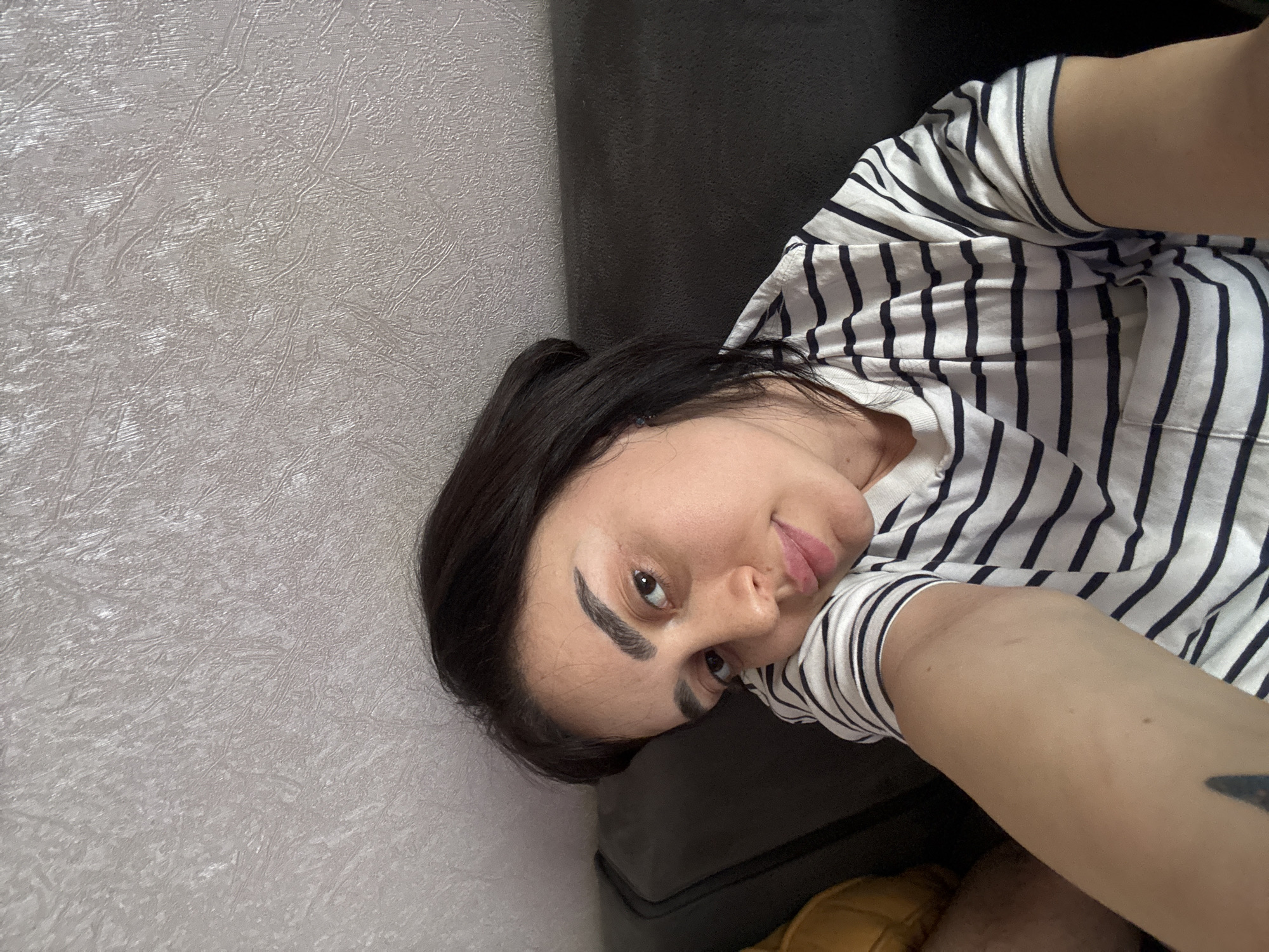 A woman with dark hair, wearing a black and white striped shirt, is lying down. She is oriented at an angle, with her head towards the left side of the frame. Part of a dark-colored couch is visible behind her head and shoulder. The background consists of a textured, light-colored wall. The city location in Tbilisi, Georgia cannot be determined from this image.
