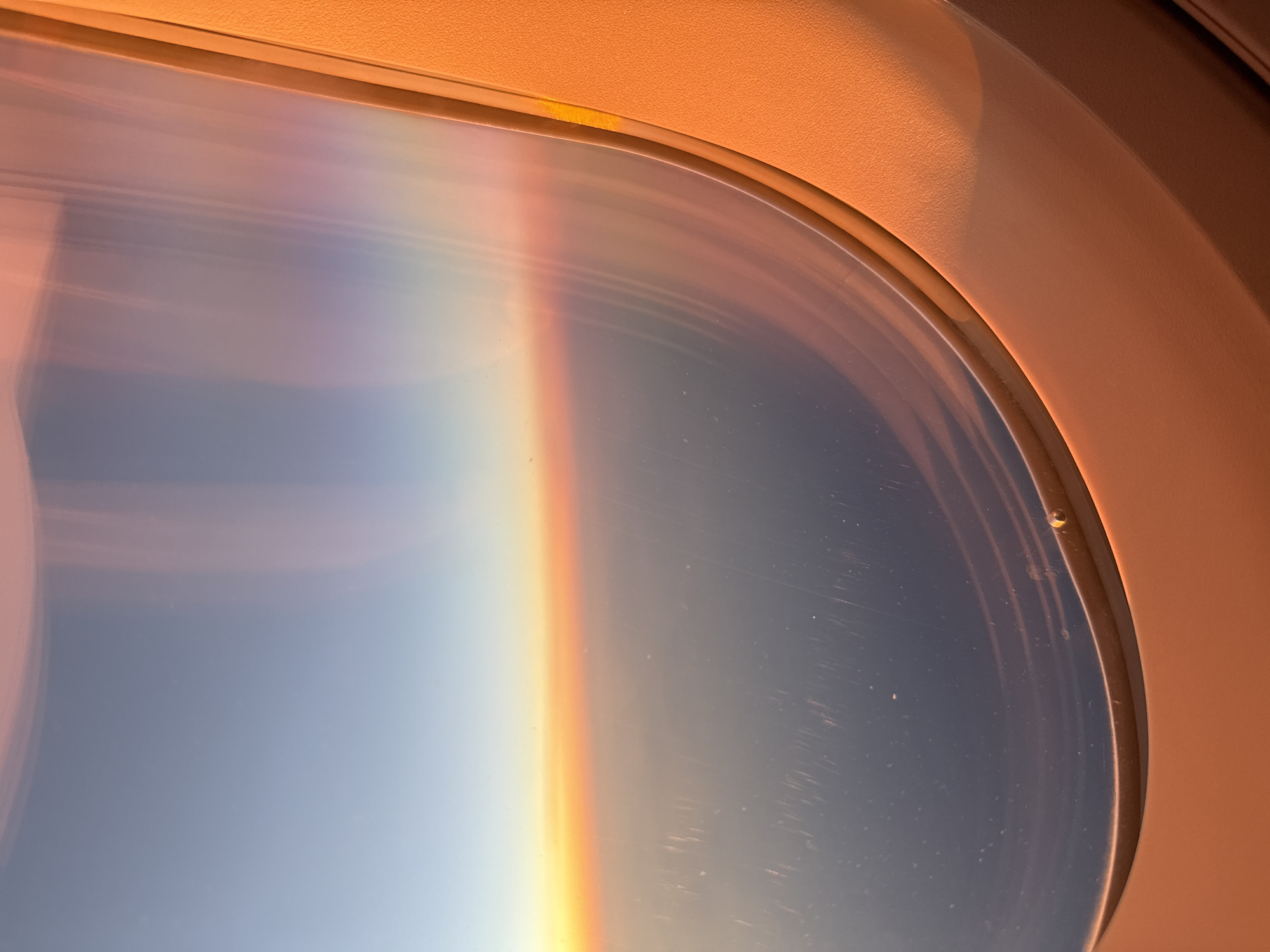 The image shows a view from an airplane window, likely at sunrise or sunset. The window frame is orange, and the view outside the window is a gradient from light blue to orange and peach. The outside view appears to be the curvature of the Earth. There is no evidence of the city of Sofia, Bulgaria. There are some scratches and imperfections visible on the window.