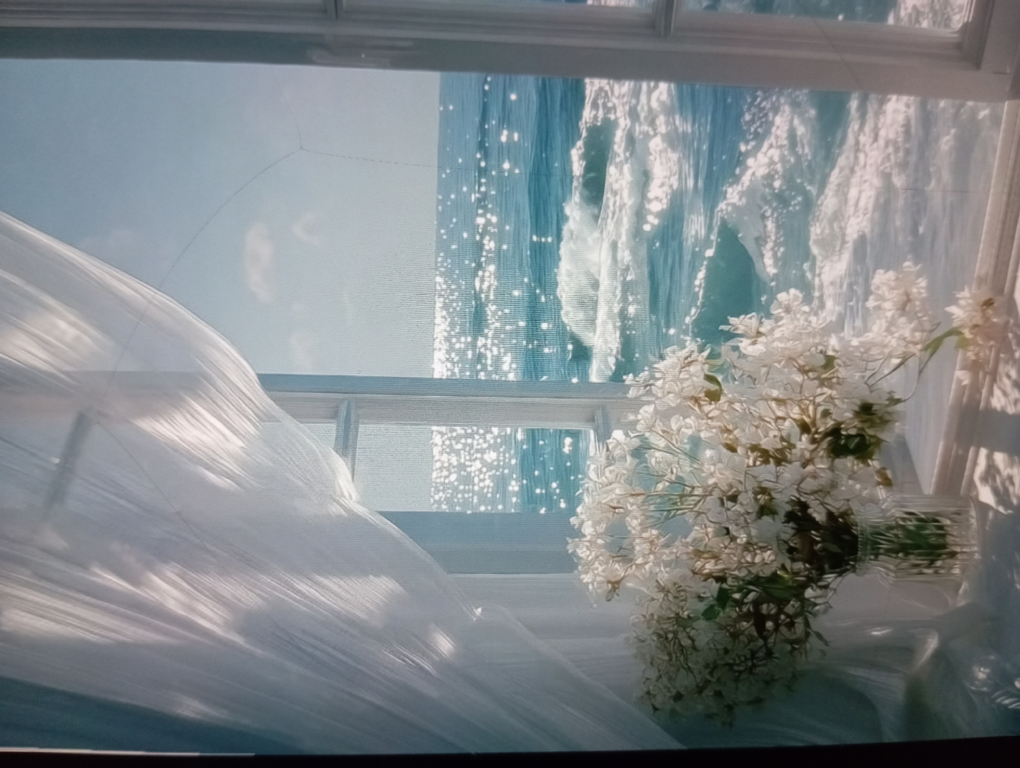 A photograph shows a view through a window. The window frame is white and divides the scene into panes. On the right side of the window sill, a vase of white flowers is visible. The flowers are in full bloom and occupy a significant portion of the lower right of the frame. A white, sheer curtain is draped across the left side of the window, billowing inwards.
Outside the window, the ocean is visible under a bright sky. The water is light blue and the sunlight reflects off the water’s surface, creating shimmering effects. The waves are clearly defined, suggesting movement or wind.
The location of Odesa, Ukraine is not discernible from the image. The scene is a generic ocean view and the photograph lacks specific landmarks or elements that would confirm this location.