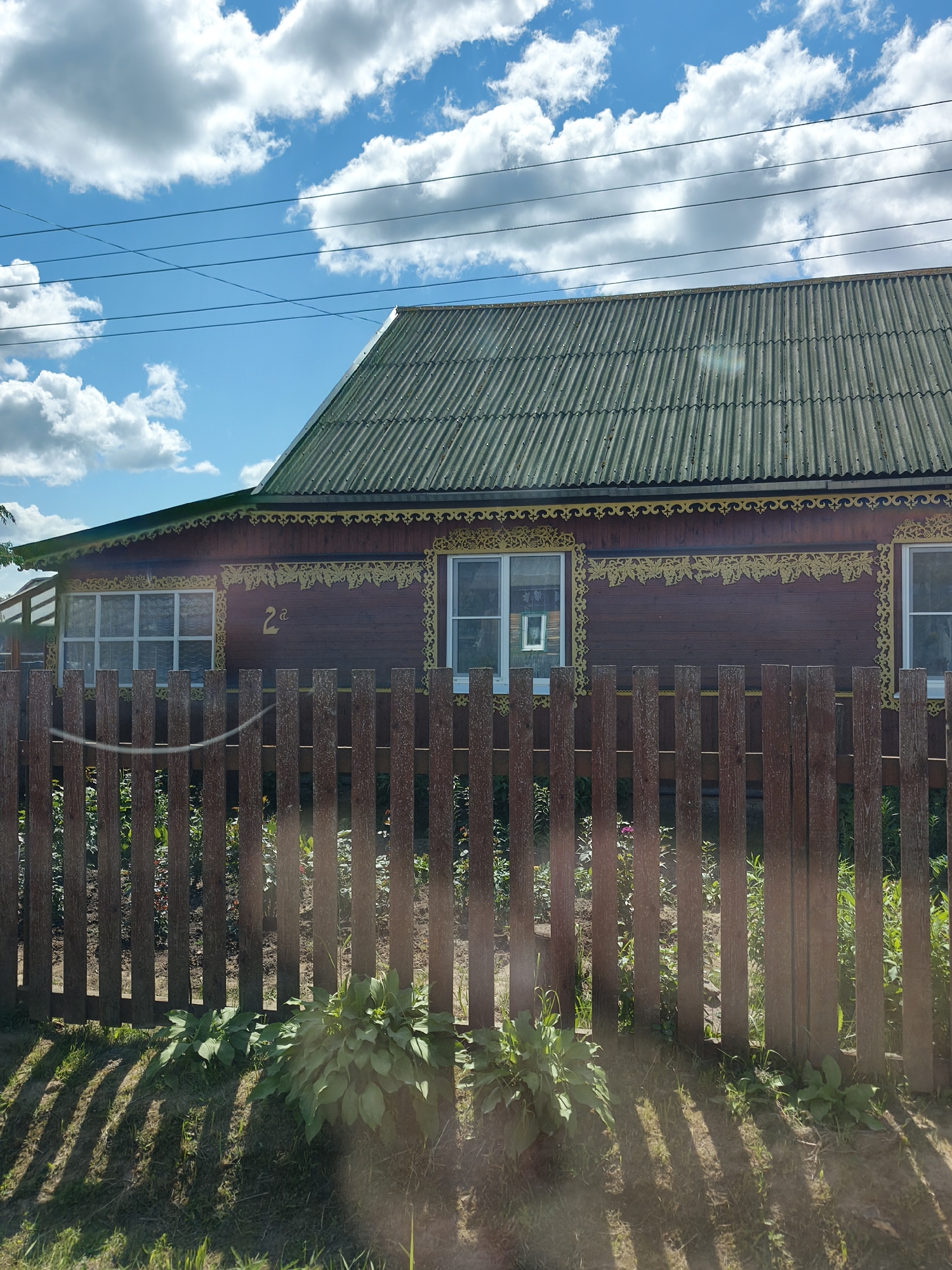 A weathered, dark brown wooden house with ornate yellow trim sits behind a vertical wooden plank fence. The house has a green corrugated roof and multiple windows. One window displays a framed picture. The number "2" followed by a Cyrillic letter "a" is painted on the house's facade between two windows. In front of the fence, lush green plants grow. The scene is illuminated by sunlight, casting shadows from the fence onto the ground. The sky above is blue with scattered white clouds, and power lines are visible against the sky. This image was taken in Kady, Russia.