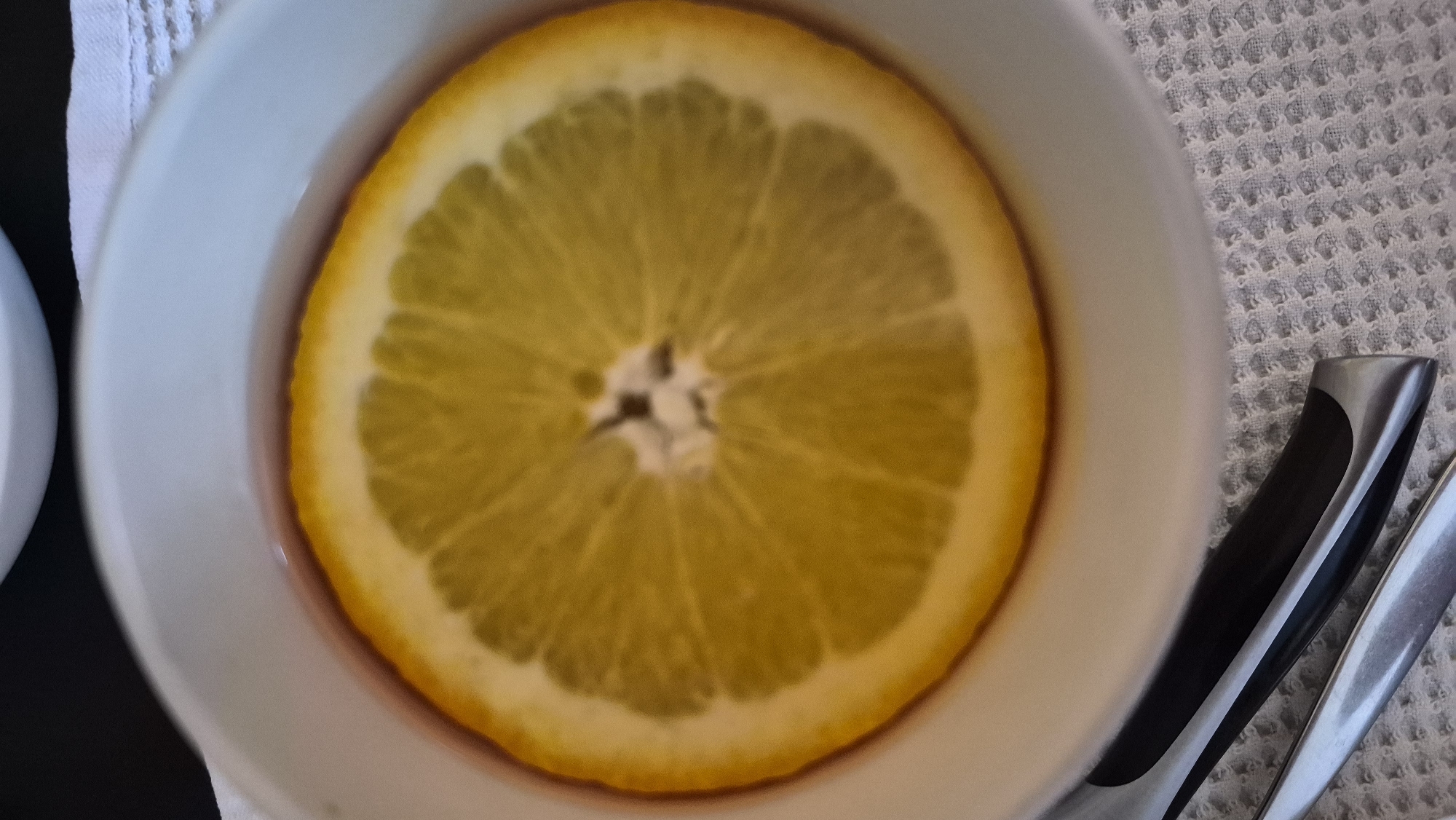 The image shows a close-up of a white bowl containing a liquid with a slice of orange floating in it. The orange slice is bright yellow with visible segments and a central core. The liquid appears to be reddish-brown. The bowl is placed on a textured white cloth. In the foreground, parts of two utensils, likely a knife and a fork, are visible, resting on the cloth. The background is dark and out of focus, with a hint of another white object above the bowl. The scene is likely indoors, possibly in a kitchen or dining area. The image does not contain any information that could place it in the city of Krauja, Latvia.