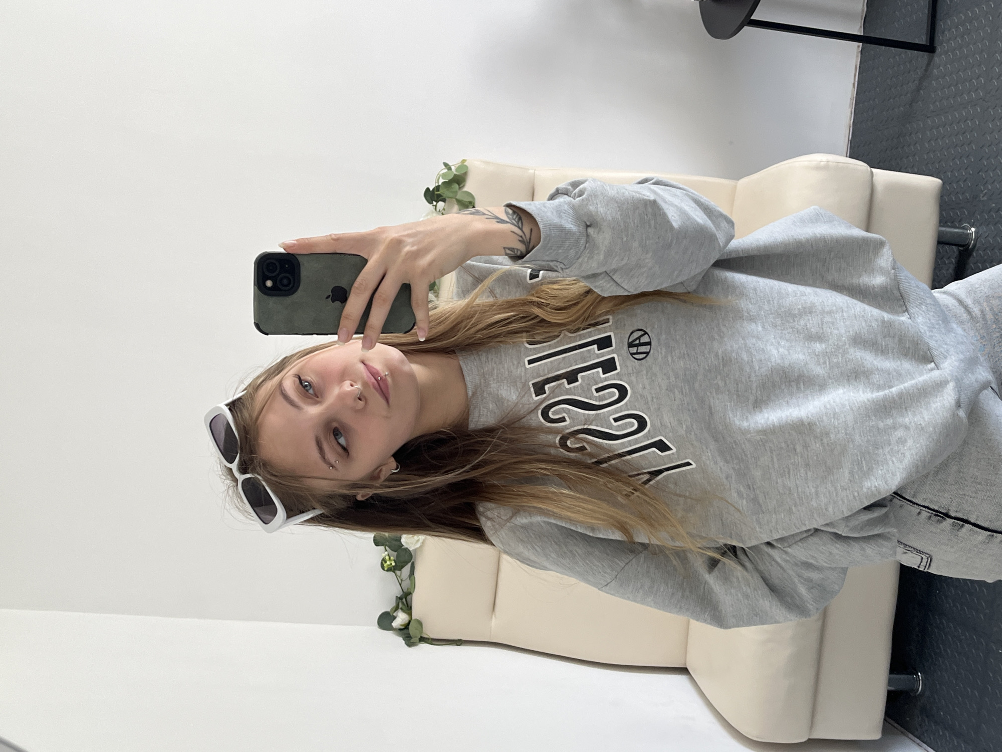A fair-skinned person with blonde hair is taking a selfie indoors. The person is wearing a grey sweatshirt with black and white lettering and white sunglasses. They are holding an iPhone with a dark green case. The background includes a cream-colored loveseat, white walls, and a black metal table. There are also decorative green vines visible. The location is Фоки, Russia.