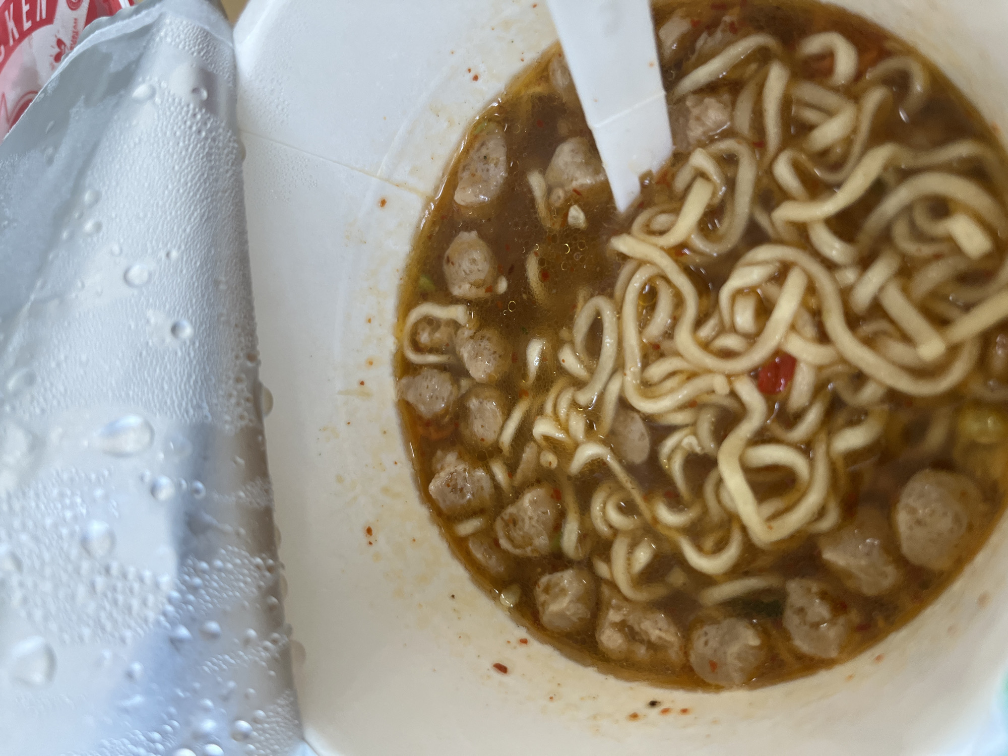 A close-up, overhead shot captures a white cup of instant noodles with meatballs and a white plastic spoon. The lid of the cup is partially open, revealing condensation droplets on its underside. The noodles are cooked and submerged in a reddish-brown broth, with small, spherical meatballs scattered throughout. The cup appears to be on a table in Kel. Pasangkayu, Indonesia.