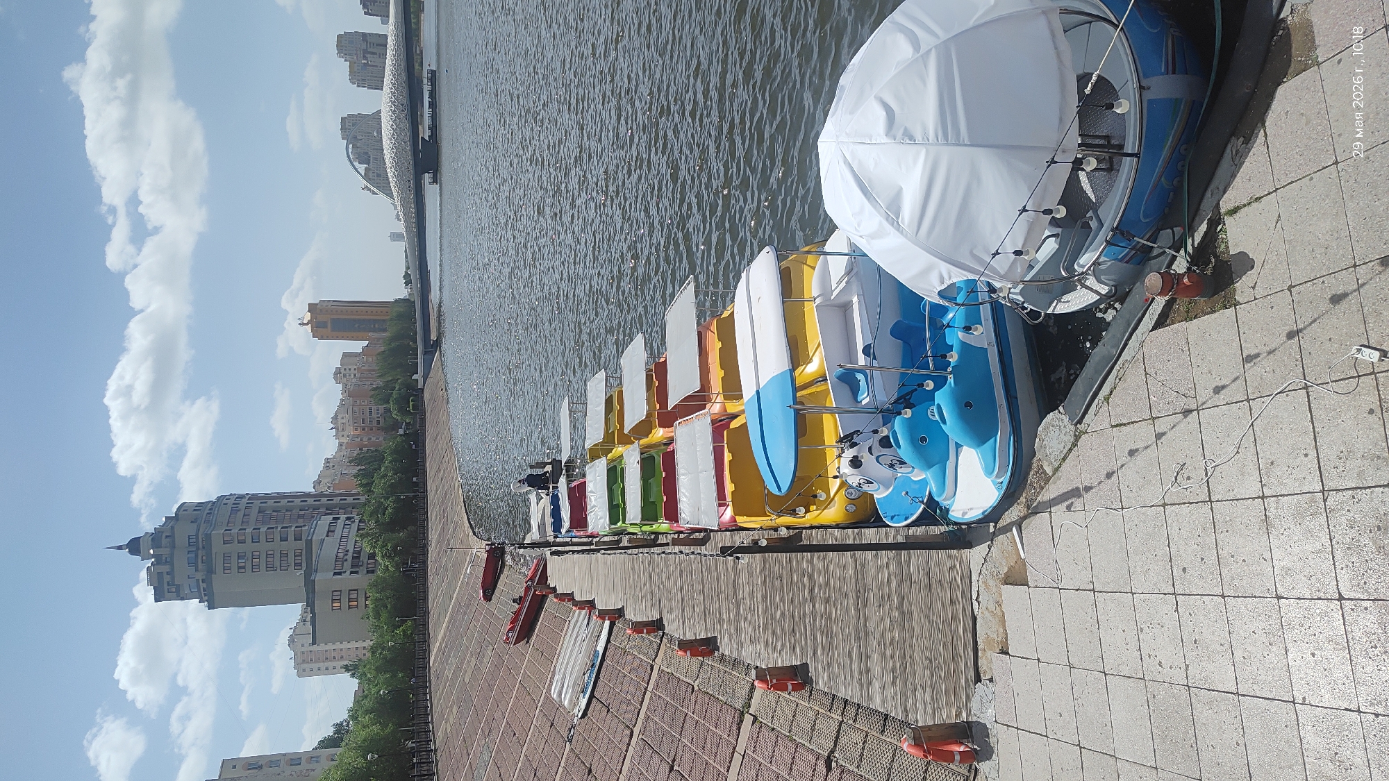 The image depicts a row of colorful pedal boats docked along a waterfront in Astana, Kazakhstan. The boats, painted in red, green, yellow, white, and blue, are neatly aligned and ready for rental. Some boats feature white canopies for sun protection. The waterfront is paved with wooden planks and brick pavers. The water itself is calm, reflecting the sky. In the background, urban buildings including high-rise apartments are visible against a blue sky with puffy white clouds.