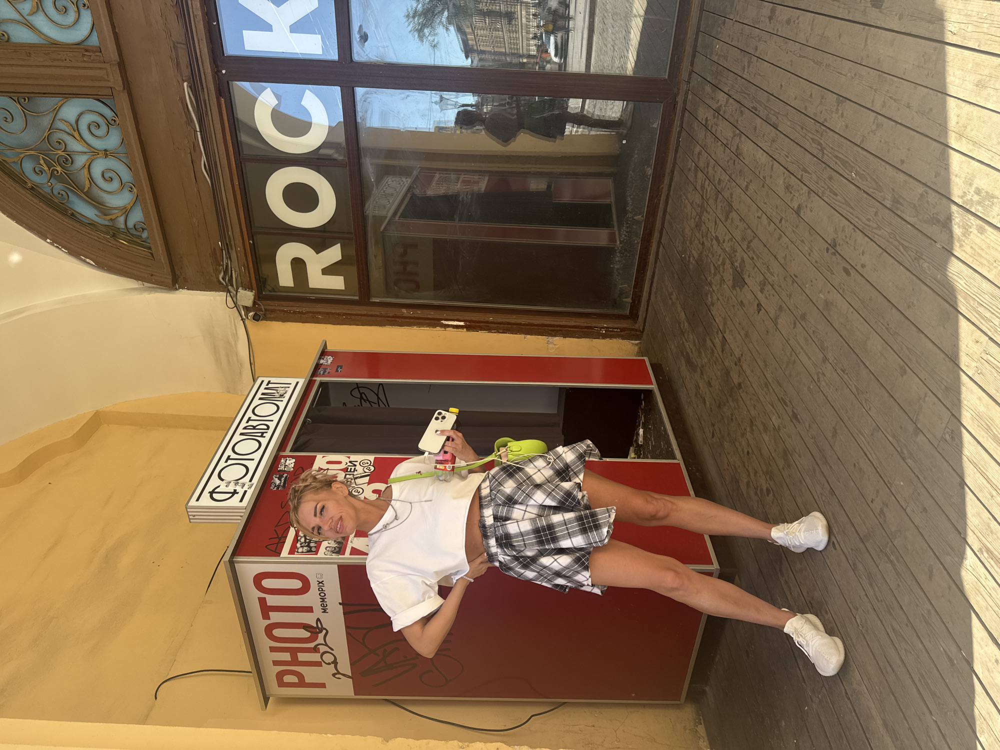 A woman with blonde hair is posing next to a photo booth that is labeled "PHOTO" and "FOTOAVTOMAT". She is wearing a white crop top and a black and white plaid skirt. She is holding a smartphone and has a green purse slung over her shoulder. The photo booth is red and white and appears to be in an outdoor setting, possibly a street in Saint Petersburg, Russia, as indicated by the Cyrillic text. The background shows a wooden walkway and a building with a large window displaying the word "ROCK" in large white letters. The scene suggests a casual, sunny day. The woman has a cheerful expression on her face.