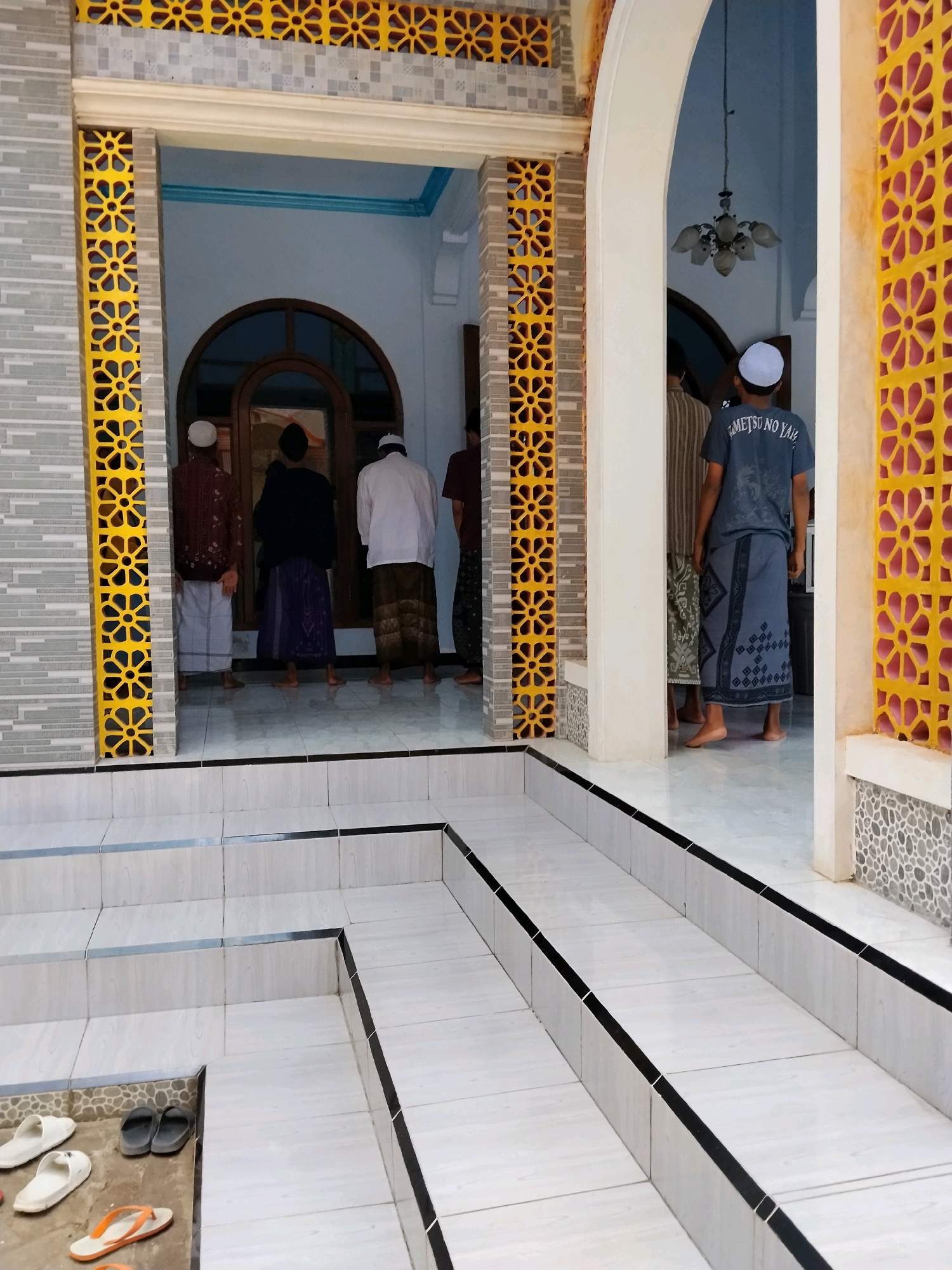 The image depicts an exterior view of a building with an arched entrance, possibly a mosque, in Tlogosari, Indonesia. Several men are standing in two groups, facing away from the camera. Some are wearing white caps and sarongs, while others are in t-shirts and sarongs. The entrance is flanked by decorative yellow and grey tiled pillars. In the foreground, a set of white tiled stairs leads up to the entrance. White and black flip-flops are visible on the lower steps. The lighting suggests it is daytime, with some shadows indicating the direction of the light source.
