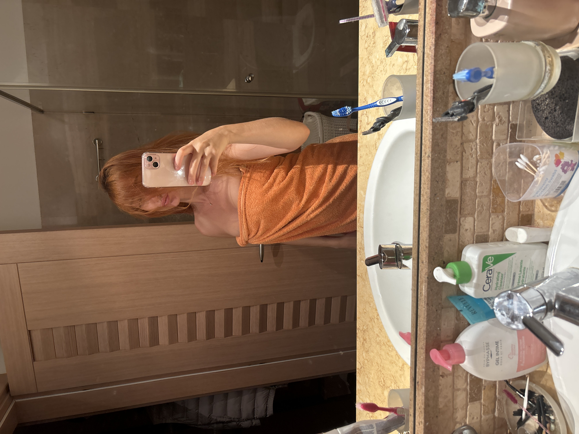 A person with red hair, wearing an orange towel wrapped around their body, is taking a mirror selfie in a bathroom. They are holding a pink smartphone in their right hand, angled to capture their reflection. The bathroom appears to have a marble countertop with various toiletries, including toothbrushes, a CeraVe cleanser, and other cosmetic items. In the background, a wooden cabinet door and a glass shower enclosure are visible. The scene suggests a personal grooming moment after showering. The location is identified as Budva, Montenegro.