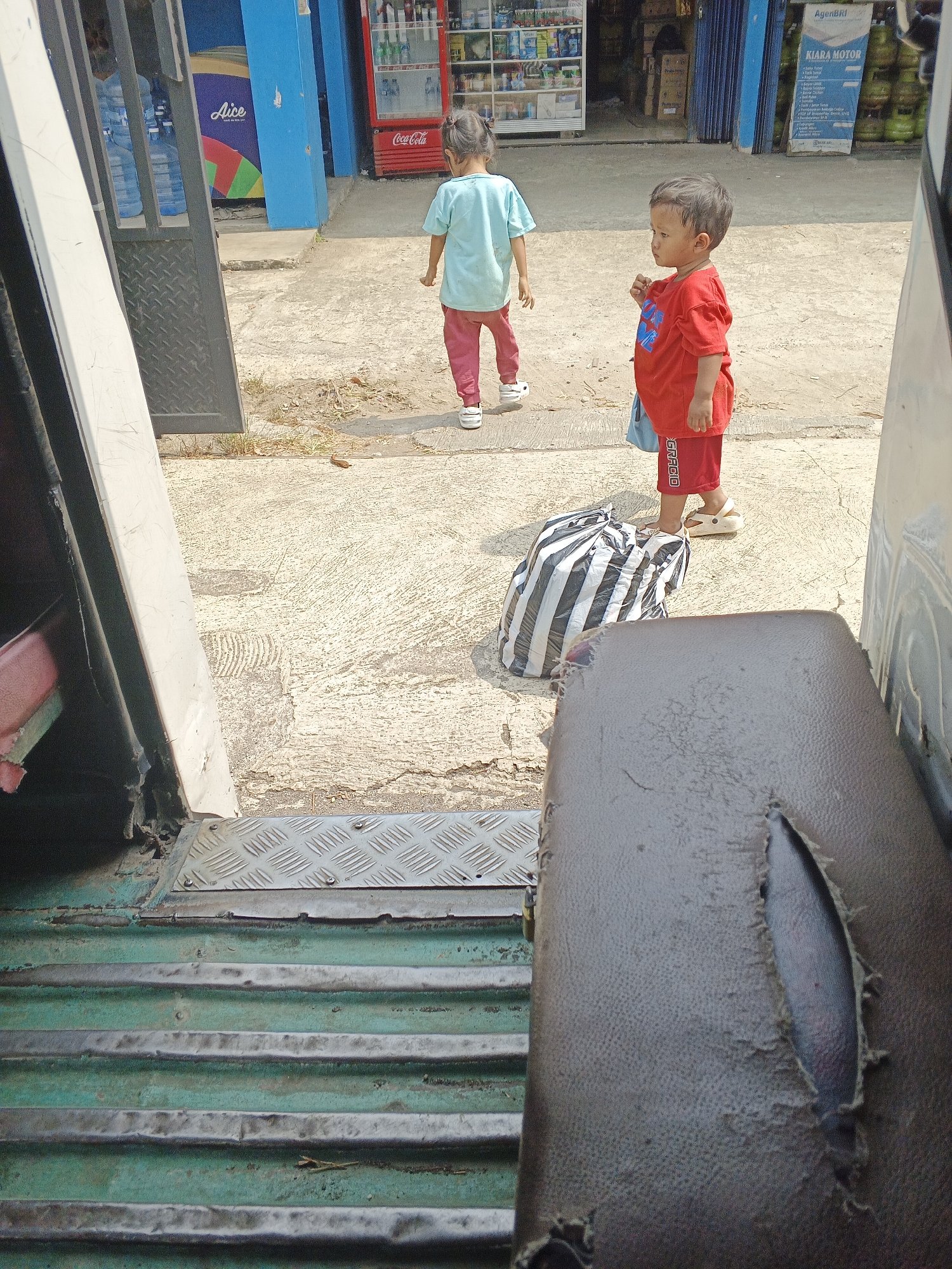 Two young children, a boy and a girl, are standing on a concrete sidewalk outside a row of shops. The girl, wearing a light blue t-shirt and pink pants, is facing away from the camera and walking towards a store with a red Coca-Cola refrigerator visible. The boy, in a red t-shirt and red shorts, is facing the camera and appears to be holding something in his right hand. He is wearing light-colored sandals. Between the children, there is a large black and white striped bag. In the foreground, a damaged car seat is visible. The environment suggests a commercial area in Warungkiara, Indonesia, with storefronts and merchandise displayed. The lighting indicates it is daytime.