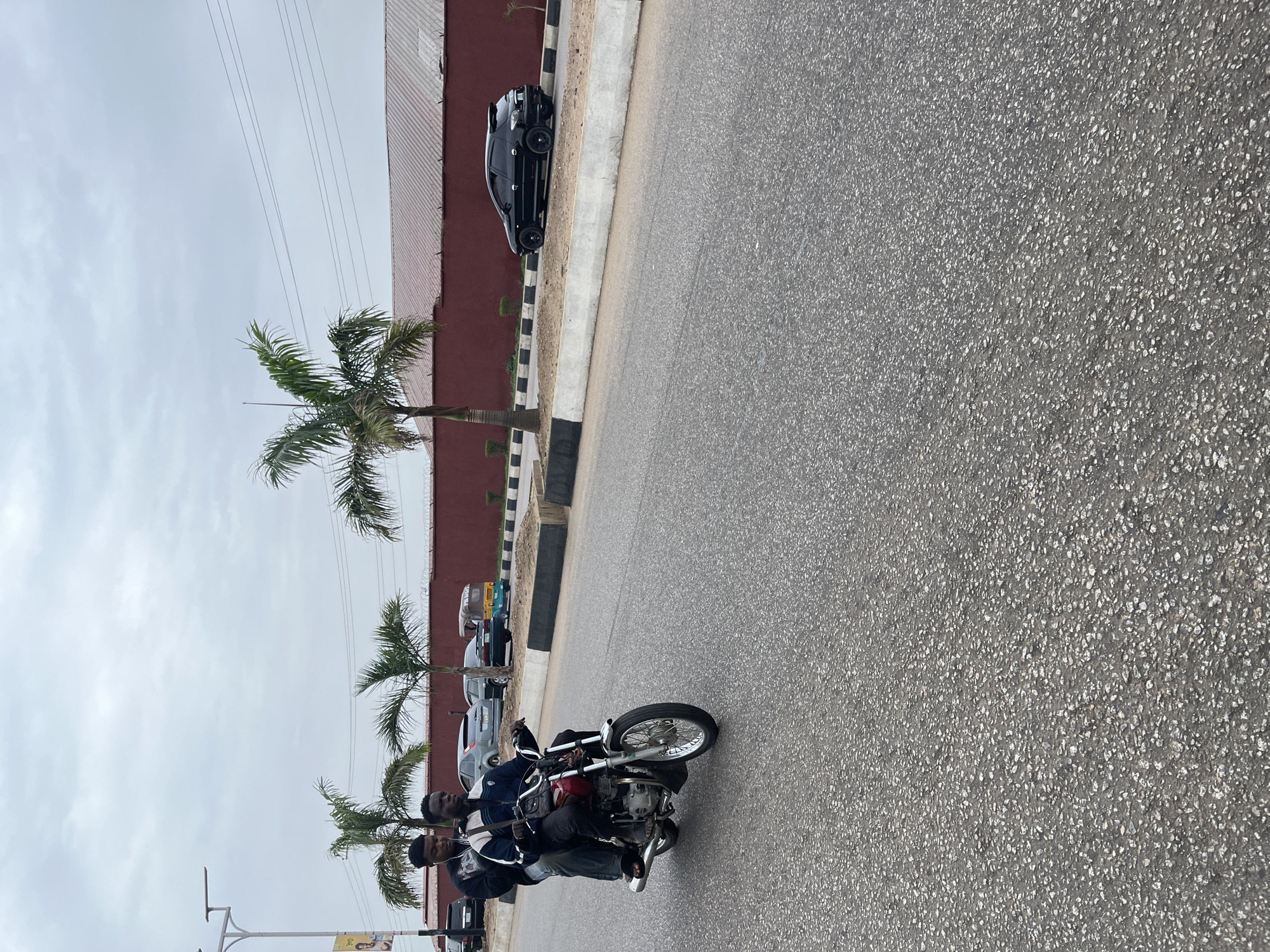 Two young Black men are riding a motorcycle on a paved road in Jos, Nigeria. The driver, in the front, is wearing a blue and white jacket and black pants. The passenger, behind him, is wearing a black hooded jacket and blue jeans. The motorcycle has a red fuel tank and a silver exhaust pipe. They are on the right side of the road, and the motorcycle is facing away from the camera.
In the background, there is a long, maroon-colored wall with a barbed wire fence on top. Several palm trees are planted along the wall. To the left of the wall, there are other vehicles parked, including a black car, a silver car, and a yellow auto-rickshaw. The sky is overcast and gray. The road surface is asphalt, and there are white and black curbs along the side of the road. The overall scene suggests an urban or suburban environment.