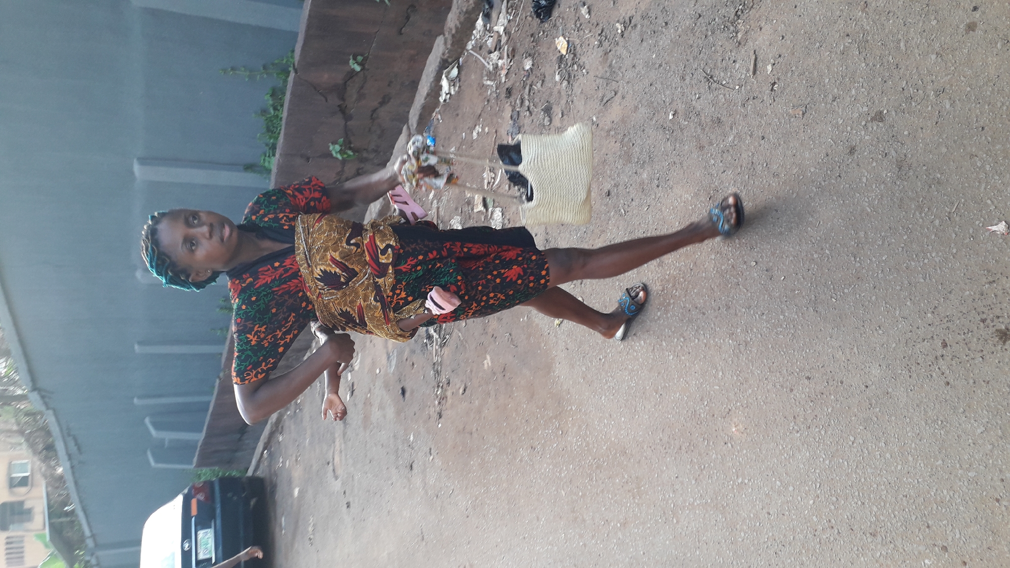 A woman stands on a road in Onitsha, Nigeria. She is carrying a baby wrapped to her front with a patterned cloth, and she also has a straw bag over her shoulder. She is wearing a printed dress and sandals. The road appears to be dirt and pavement mixed. Buildings and vegetation are visible in the background, as is a car.