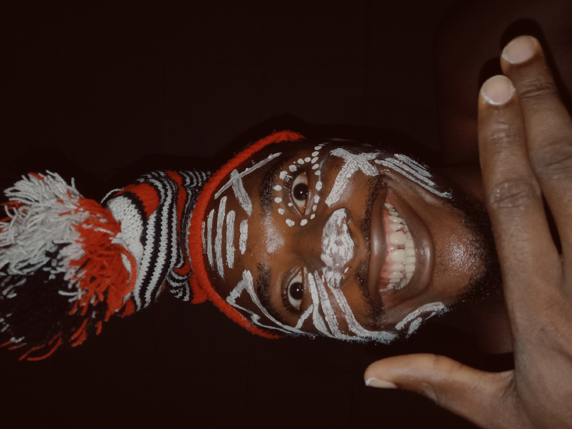 A dark-skinned person with white paint markings on their face and a knitted hat with red, white, and black stripes is smiling. The person's face is angled, with a hand partially covering the right side of the face. The background is dark and indistinct. The location is Unknown, Unknown.