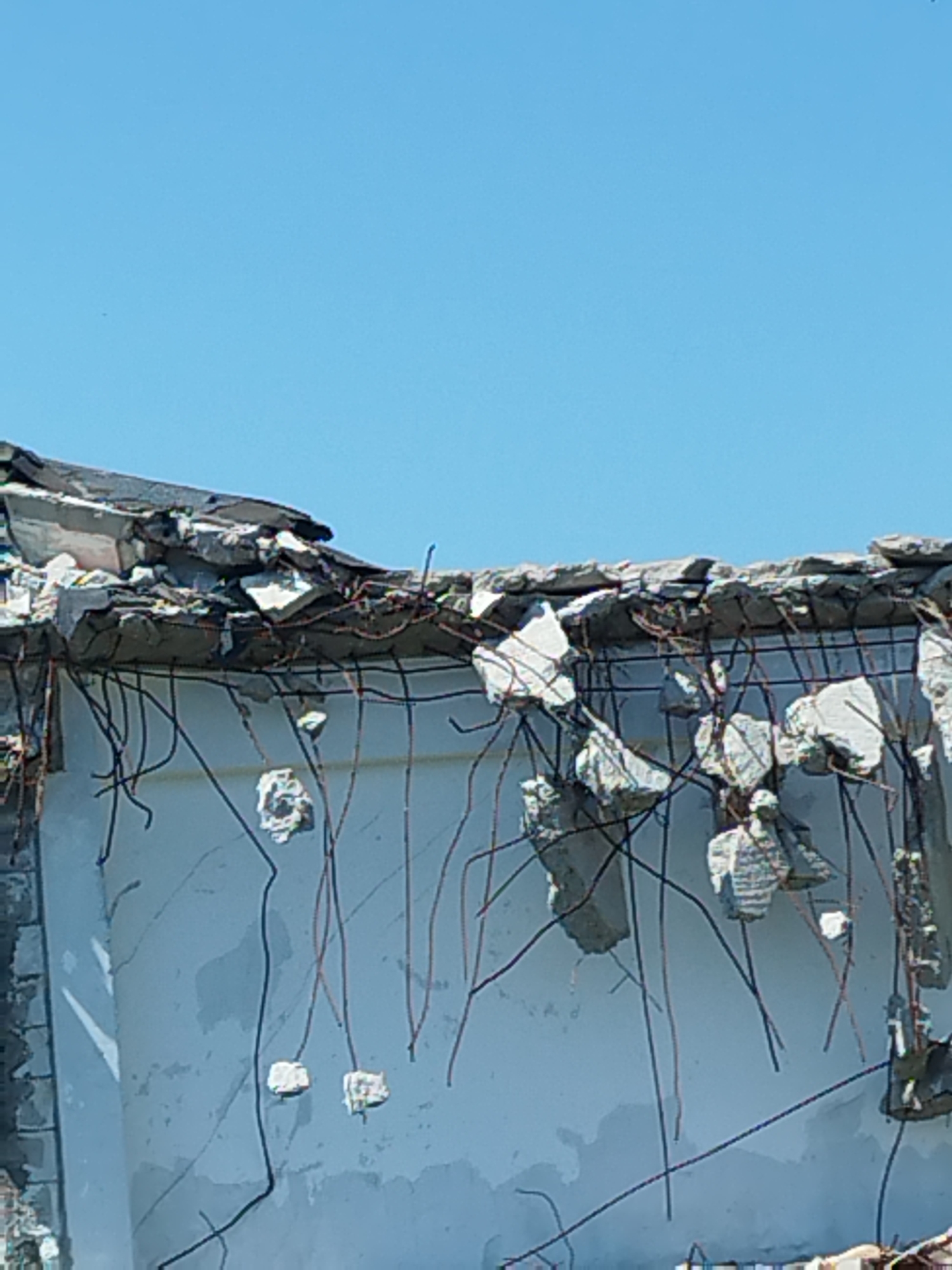 The image captures the upper section of a partially demolished building against a clear blue sky. The exposed concrete structure reveals rebar protrusions and broken concrete fragments, indicating active demolition or significant structural damage. The wall below the damaged upper section appears to be a light blue or gray, with visible cracks and a somewhat rough texture. Scattered across the wall are various pieces of concrete debris, some of which are attached to dangling rebar. The overall scene suggests a construction or deconstruction site. The location is identified as Clinceni, Romania.
