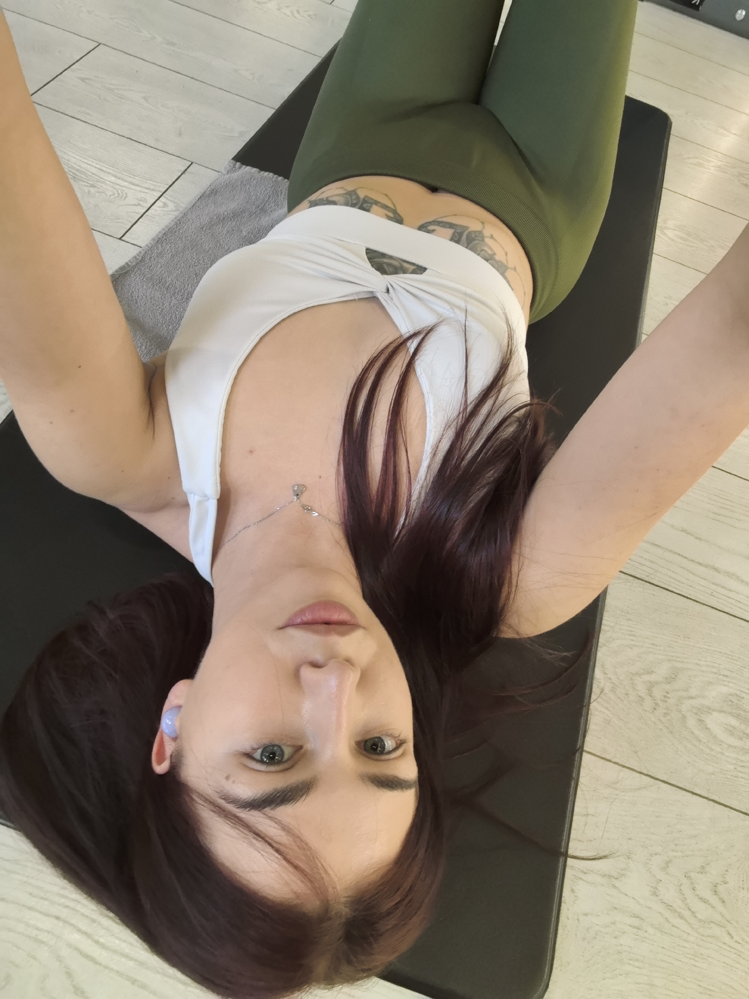 The image depicts a person lying on a black yoga mat. The person is wearing green leggings and a white top. They have dark hair, fair skin, and are wearing a blue earbud in their left ear. A gray towel is visible near the top of the mat. The floor is light wood grain. No information within the image identifies a location in Stara Zagora, Bulgaria.