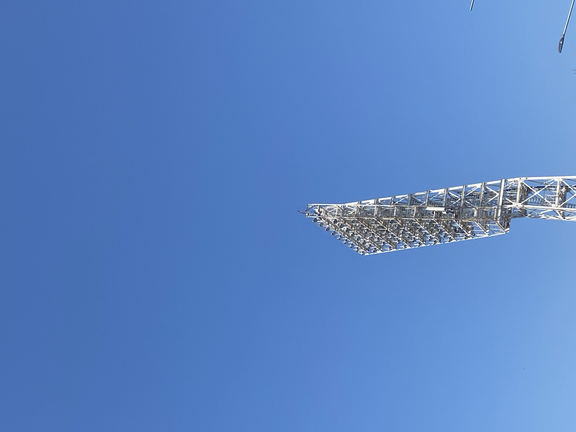 The image shows a stadium light tower under a clear blue sky. The location is Заволжье, Russia. The tower is constructed from a white metal framework and holds an array of spotlights. There are no people visible.