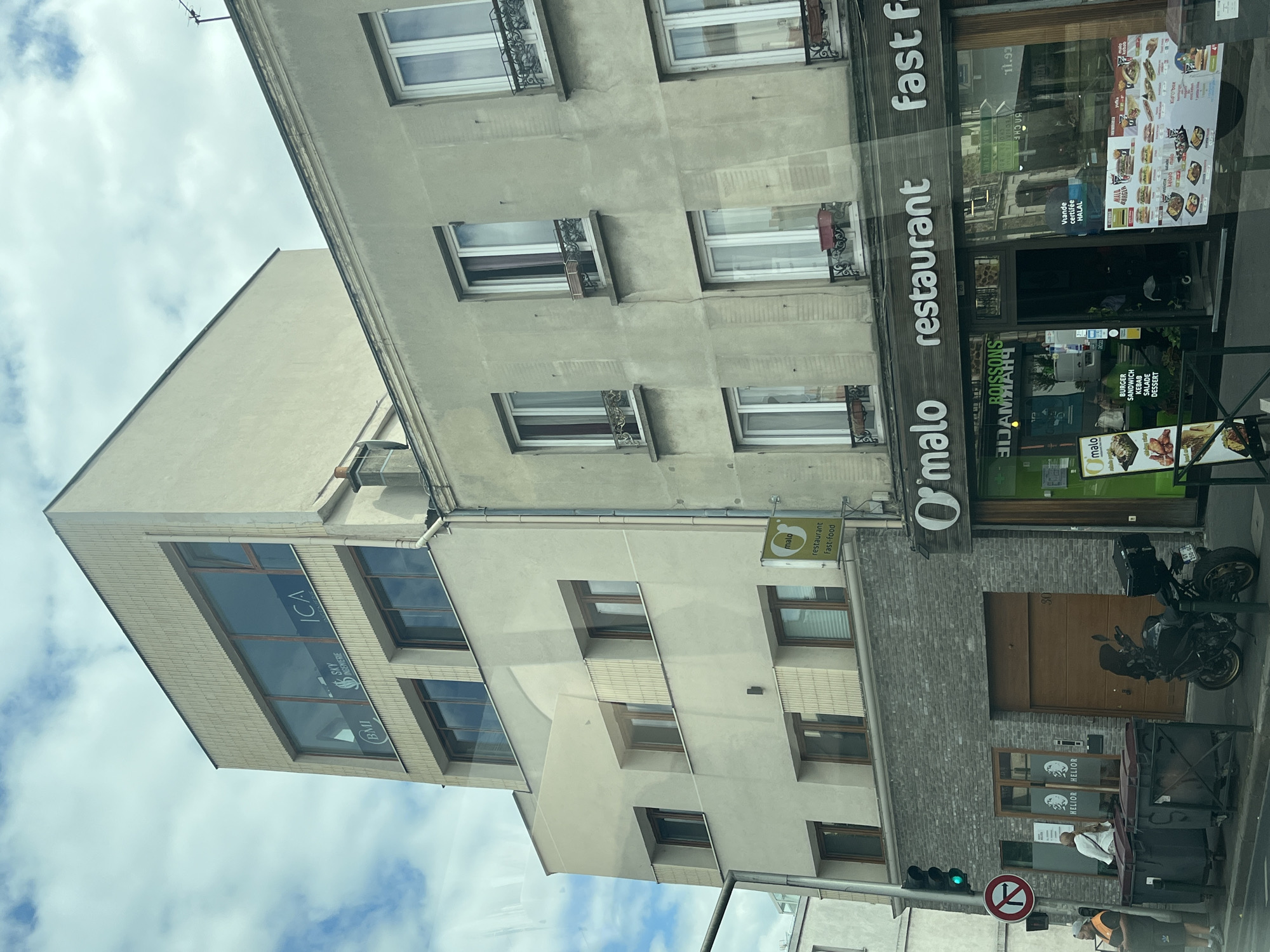 This is a street-level, eye-level, medium-range, eye-level shot of a street in Alfortville, France, on a partly cloudy day. The scene features a modern building with a grey brick lower level and a beige upper section with large windows. To the right, an older, grey building with multiple windows is visible. A sign for "O'malo restaurant fast food" is prominently displayed above the entrance of a restaurant, which features various promotional posters for food items. A green traffic light is visible on the left, along with a traffic sign indicating a no-entry zone and two large trash bins. A man in a reflective vest stands near the bins, and a black motorcycle is parked in front of a wooden garage door on the lower level of the modern building. The windows of the modern building display company names such as "ICA", "SKY INGENIERIE", and "BMI".