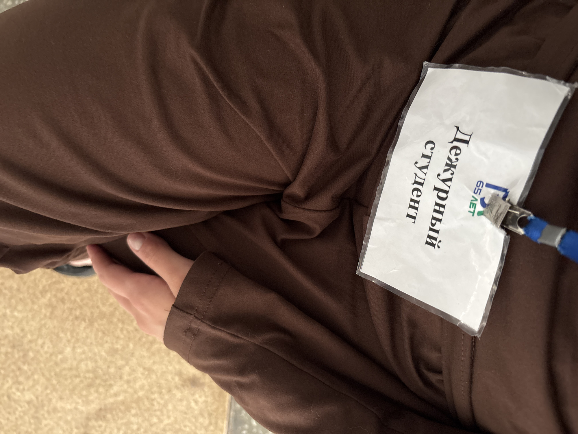 The image shows a close-up of a person's hand resting on brown fabric, likely clothing. Attached to the fabric is a white badge in a plastic sleeve. The badge contains text in Russian. The visible portion of the text reads "Дежурный студент" (Dezhurnyy student), which translates to "Duty Student". Below this, the text "65 ЛЕТ" (65 Let), meaning "65 Years", is visible along with a blue lanyard and a silver clip. The background appears to be a textured, light-colored surface, possibly carpet or a similar material. The lighting suggests an indoor setting. The image does not contain any specific landmarks or visual cues that would allow for the precise identification of a location within Belovo, Russia.