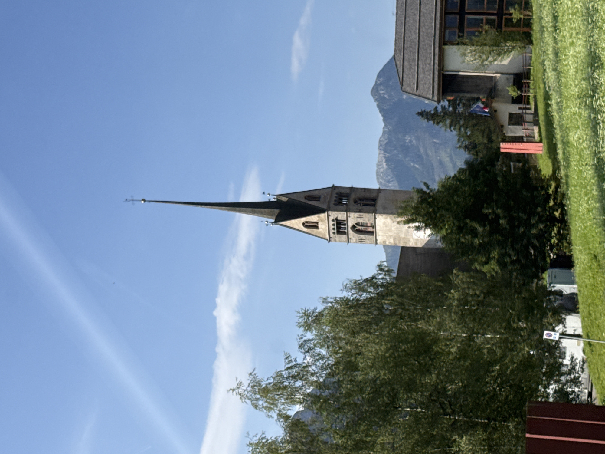 The image captures a daytime scene in San Giovanni di Fassa - Sèn Jan, Italy, dominated by a prominent church spire reaching towards a clear blue sky with wispy clouds. The spire is part of a stone church building with arched windows and decorative elements. In the foreground, lush green trees with dense foliage partially obscure the view, with a smaller building to the right of the church. This building has a dark, sloped roof and large windows. A small structure with a flag (appears to be the French flag) is situated in front of this building. In the background, a majestic mountain range with rocky peaks rises against the horizon. The ground in the foreground is covered with green grass and a scattering of small white flowers. A red vertical structure is visible on the left side of the image, and a white signpost with a symbol is planted in the grass.