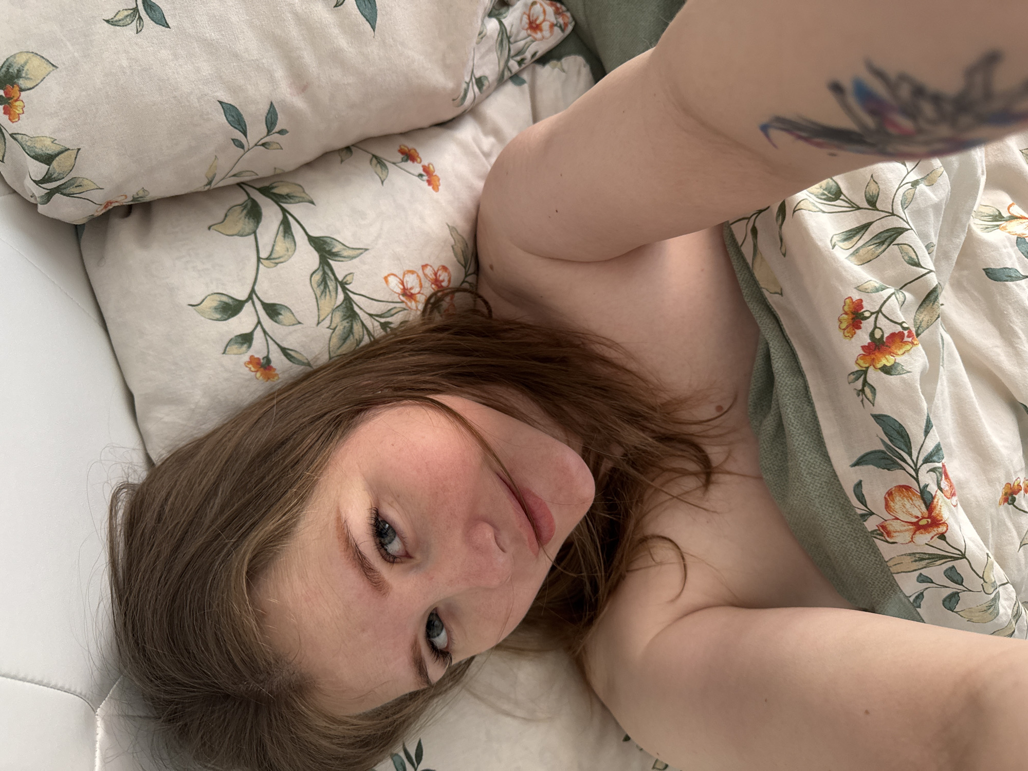 A young woman is captured in a close-up, high-angle selfie, lying comfortably on a bed. She has medium-brown hair, which frames her face, and striking blue eyes with prominent eyelashes. Her expression is soft and serene, accompanied by a gentle, slightly parted smile. Her skin is fair, with a few visible freckles.
She appears to be bare-shouldered, with her body partially covered by a duvet or blanket. This bedding features a light, off-white background adorned with a repeating pattern of green leaves and small, vibrant orange and red flowers. A plain light green or teal fabric is visible as a trim or second layer next to the floral pattern. Her right arm, extended to hold the camera for the selfie, is visible in the upper right portion of the frame, revealing a colorful, blurred tattoo on her upper arm that includes shades of blue, red, and black.
Behind her head, two pillows matching the floral and leaf pattern of the duvet are visible. To the left of her head, a white, possibly upholstered or padded surface, likely a headboard or part of the bed frame, is seen. The lighting in the image is soft and even, suggesting an indoor setting, possibly during the morning or early afternoon in Perm, Russia. The overall atmosphere conveyed is one of relaxation and calm. There is no visible text in the image.