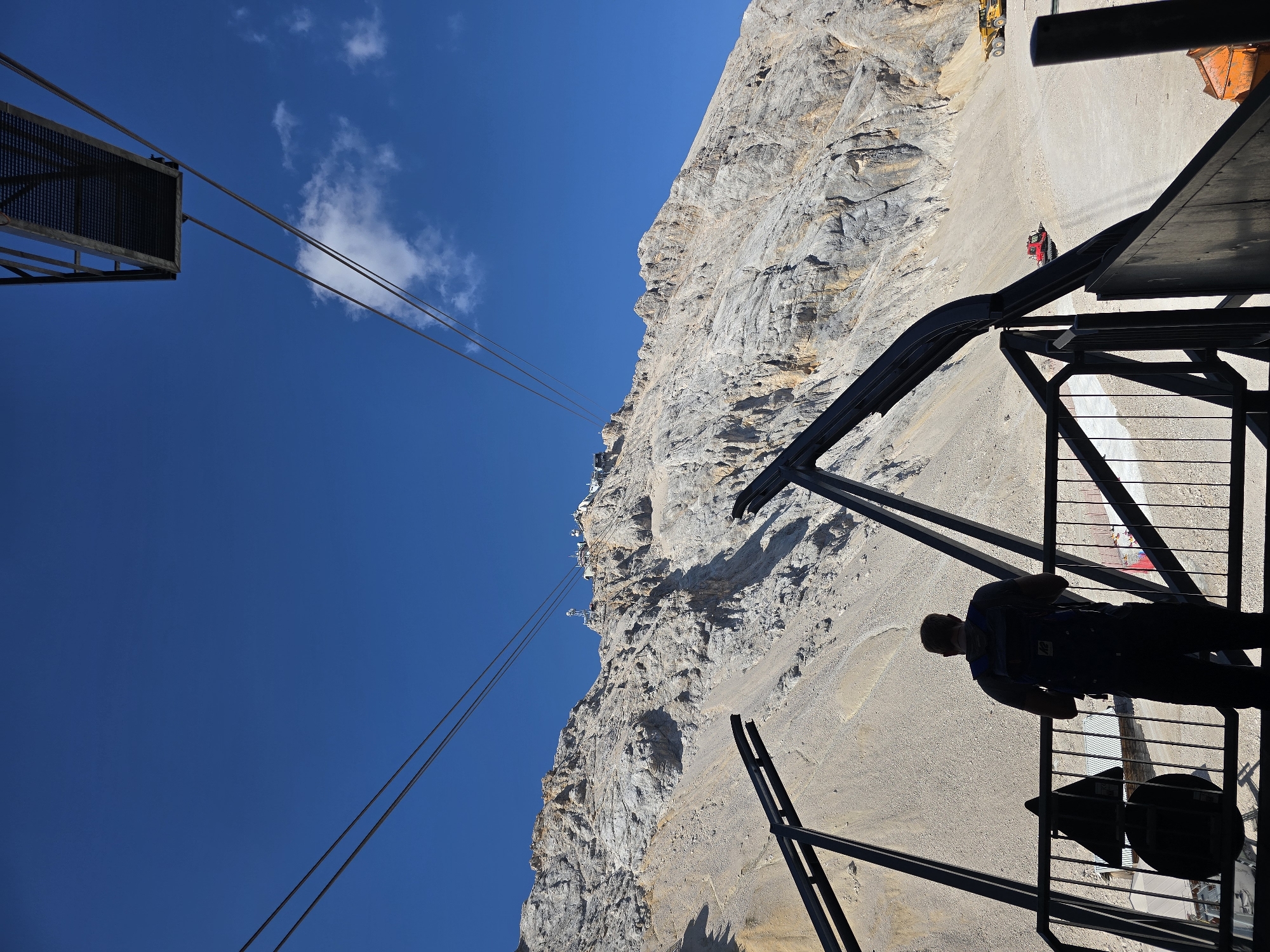 The image depicts a scenic view of a mountain landscape with a clear blue sky. In the foreground, a person wearing a backpack stands on a platform, looking towards the mountain. The platform appears to be part of a cable car station, with cables stretching across the frame. The mountain itself is rugged and rocky, with some structures visible at the summit. In the mid-ground, there are some construction vehicles, including a red snow groomer and a yellow dump truck, suggesting ongoing work or maintenance on the mountain. The overall impression is one of a tourist destination with outdoor activities and infrastructure for accessing high altitudes, likely in the Bavarian Alps near Garmisch-Partenkirchen, Germany.