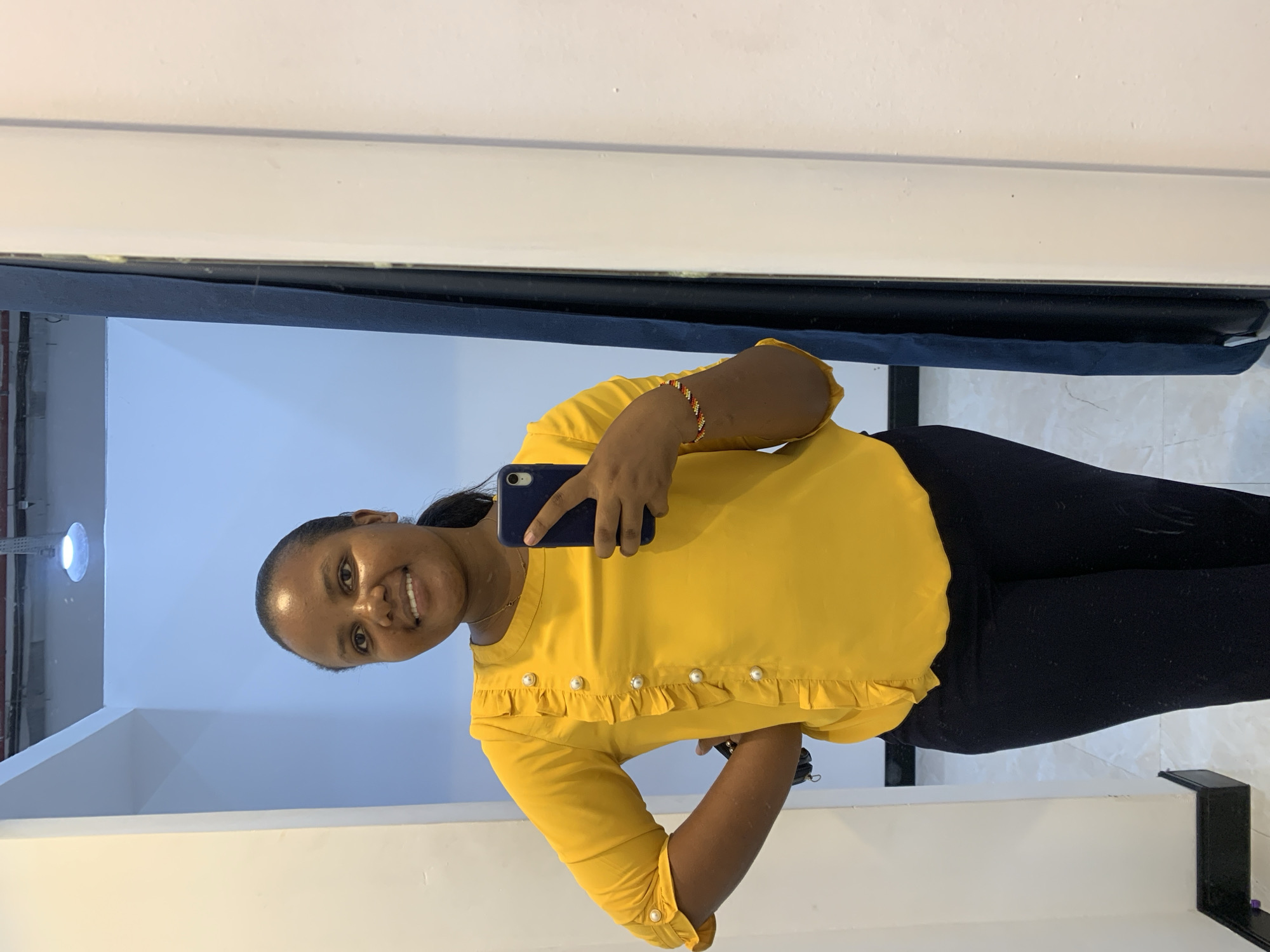 A woman is taking a selfie indoors. She is wearing a yellow blouse with pearl embellishments and black leggings. She is holding a blue smartphone. The background includes a blue curtain, white walls, and a tiled floor. The image appears to be a reflection, suggesting it was taken in front of a mirror. There is no information about the city of Dar es-Salaam, Tanzania, so location specifics are unknown.