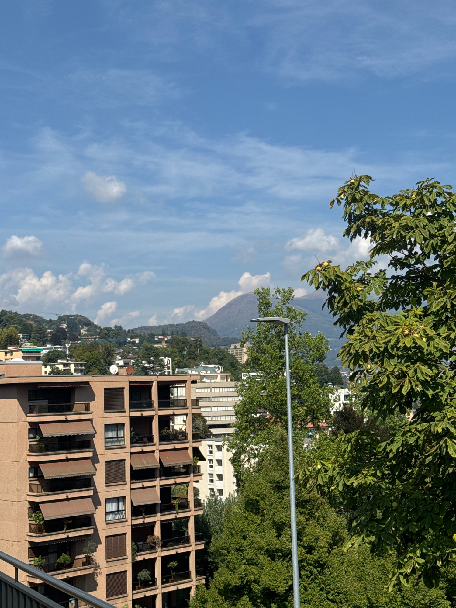 The image depicts an urban landscape in Lugano, Switzerland, under a bright blue sky with scattered white clouds. In the foreground, a multi-story building with balconies and awnings is visible on the left. To the right, a tall, leafy tree with some round fruits can be seen. A street lamp stands between the building and the tree. In the background, more buildings, lush green hills, and a distant mountain peak are present. The scene appears to be a daytime view from an elevated position.