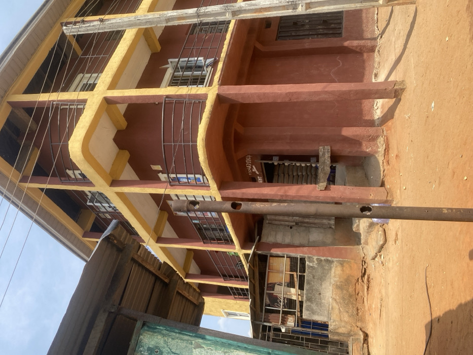 The scene depicts an exterior view of a three-story building with reddish-brown walls and prominent yellow-painted beams and balcony railings, situated in Iyiowa Odekpa, Nigeria. The building features multiple windows, some open, and balconies with dark metal railings on each floor. A ground-level entrance displays a partially visible sign with text "MENT" or "CEMENT" and phone numbers "0704307064", beneath which stacked white sacks are visible, indicating a commercial establishment. To the left, a single-story structure with a corrugated metal roof and partially green-painted walls is visible, including a "Coca-Cola" sign. The ground is unpaved, consisting of dry, reddish-brown earth. A weathered metal pole with circular holes stands vertically in the mid-ground. The sky is clear blue. No individuals are present, and no actions or interactions are depicted.