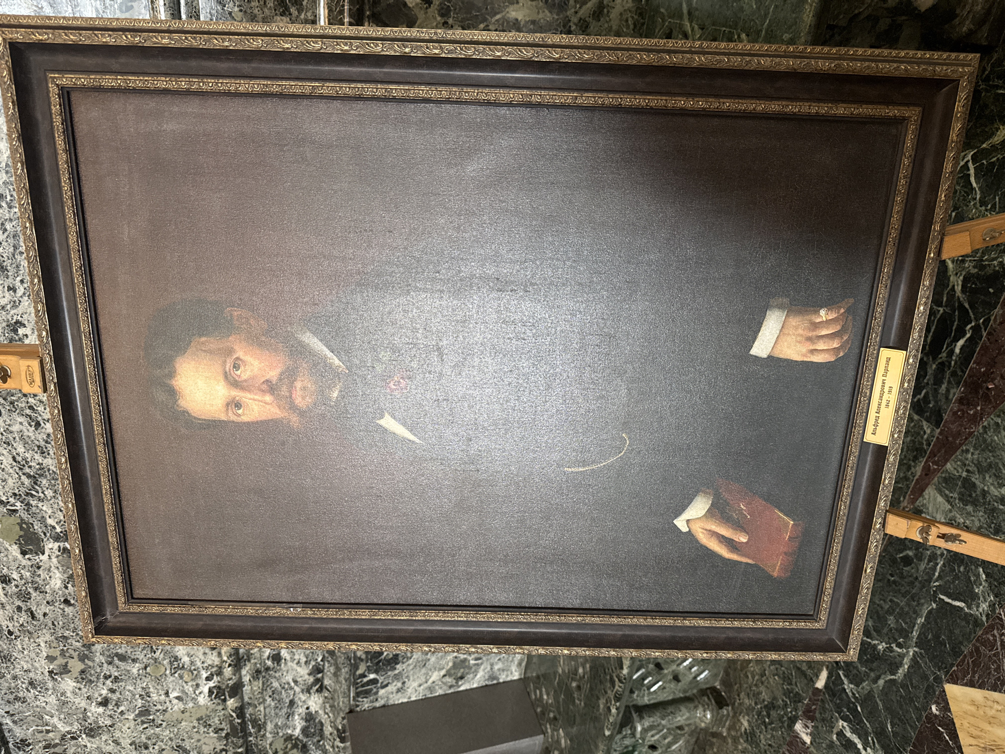 The image shows a framed portrait hanging on a wall. The portrait depicts a man with dark hair and a beard, wearing a dark suit with a white shirt and bow tie. His hands are visible, one holding a book and the other adorned with a ring. A small label on the lower right of the frame appears to contain text in Russian. The environment is a room with a marbled floor and a wall with green marble elements. The location is Saint Petersburg, Russia, given the portrait's likely Russian origin and the common use of marble in the city's historical buildings.