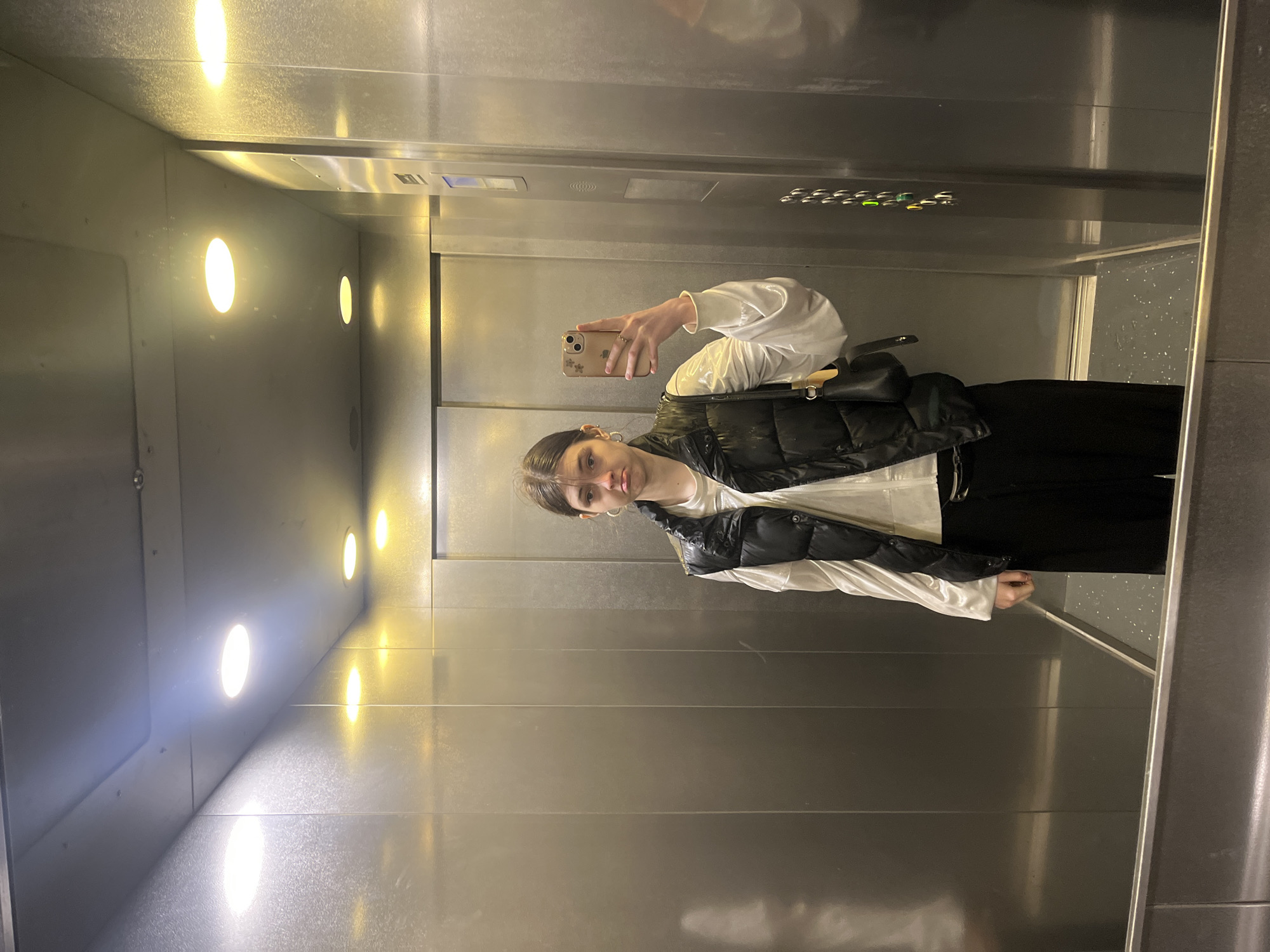 A young person is taking a selfie in an elevator. The elevator has a metal interior with a vertical row of round lights on one side. A control panel with buttons is visible near the top. The subject is wearing a white top, a black vest, and black pants. A black bag is slung over their shoulder. The person is holding a phone with a patterned case. The location cannot be determined from the image.