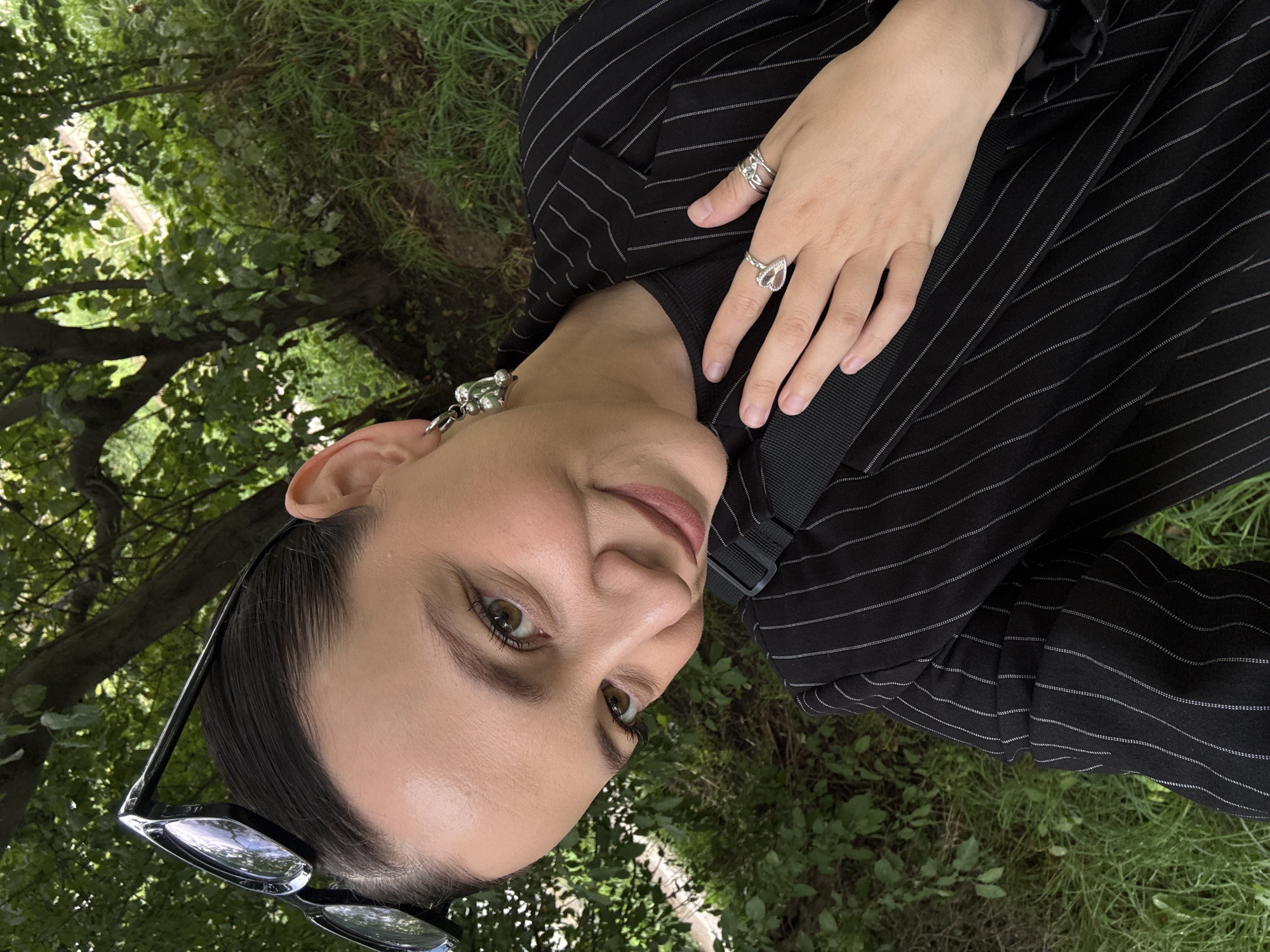 This media file shows a close-up, rotated, and slightly tilted selfie of a person outdoors. The subject is wearing a black pinstripe blazer and multiple silver rings on their right hand, including one with a teardrop-shaped stone. They are also wearing a large, silver-toned earring. The person has dark hair pulled back and is wearing makeup, including eyeliner and mascara. Their gaze is directed towards the camera.
The background consists of green grass and trees, suggesting a natural outdoor setting, possibly a park or garden. The lighting appears to be natural daylight, casting soft shadows, which, along with the lush greenery, indicates it's likely daytime during a season with foliage, such as spring or summer. The overall mood conveyed is casual and relaxed, with the person appearing content. There is no visible text in the image. The context provided suggests the location is Chaikovsky, Russia, which aligns with the natural, green environment.